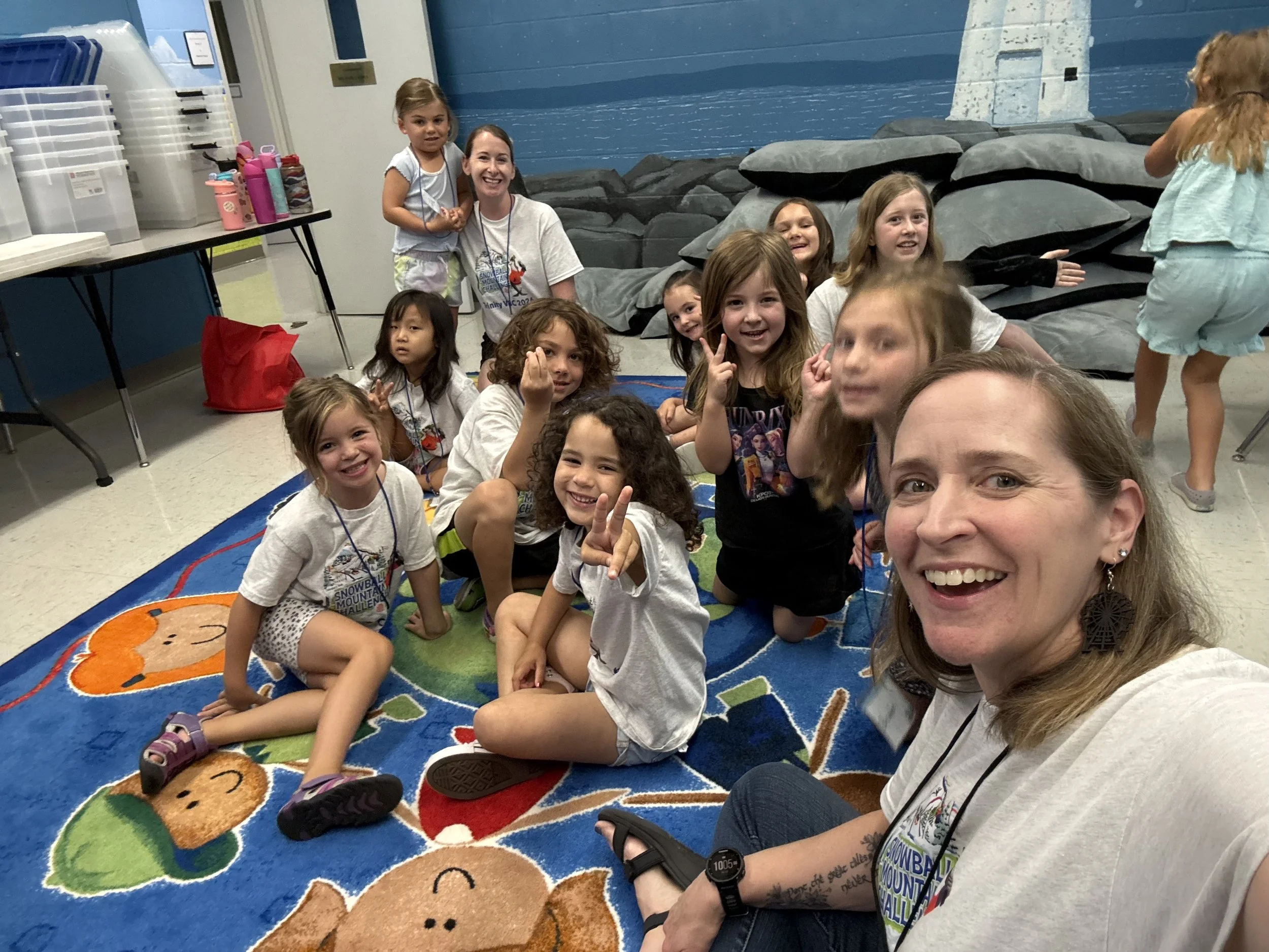A group of children and an adult woman sitting on a colorful rug with cartoon animal designs, smiling and making peace signs. In the background, a blue wall with a mural of a lighthouse and rocks, and a table with water bottles and storage containers.