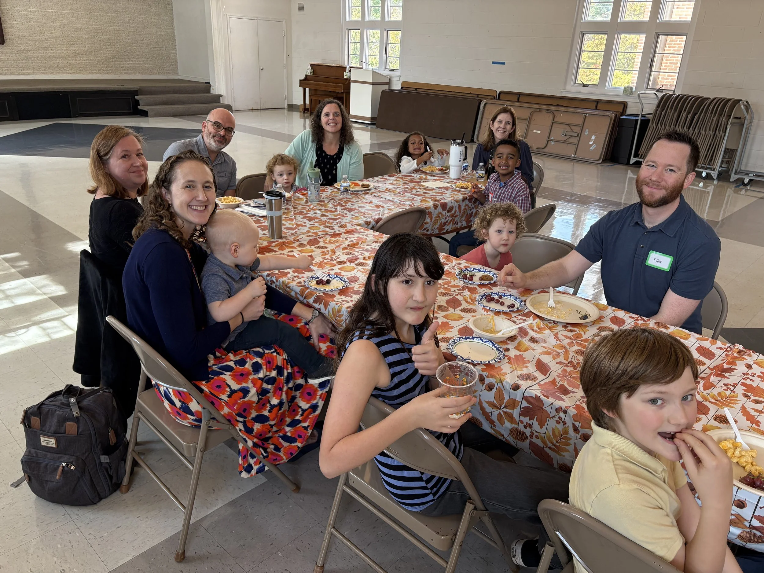 A group of children and adults sitting at a long table with a fall-themed tablecloth, enjoying a meal in a spacious room with large windows, a piano, and stacked folding tables in the background.