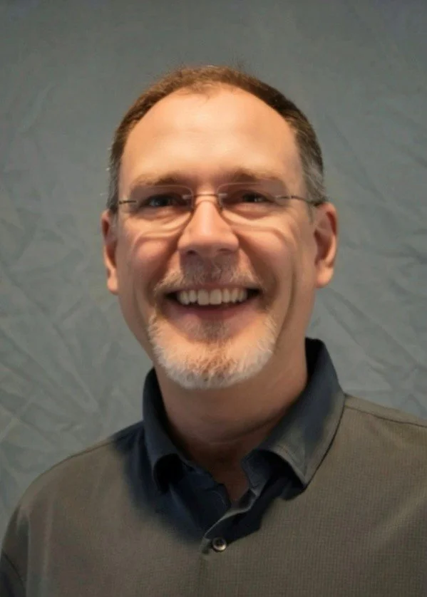 A middle-aged man with glasses, smiling broadly, dressed in a gray checkered shirt, standing indoors against a wall with subtle textured pattern.