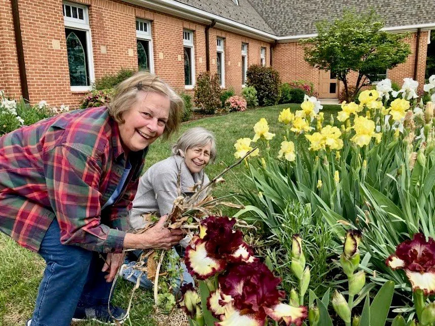 Two women planting flowers in a garden in front of a brick house with a manicured lawn and trees.