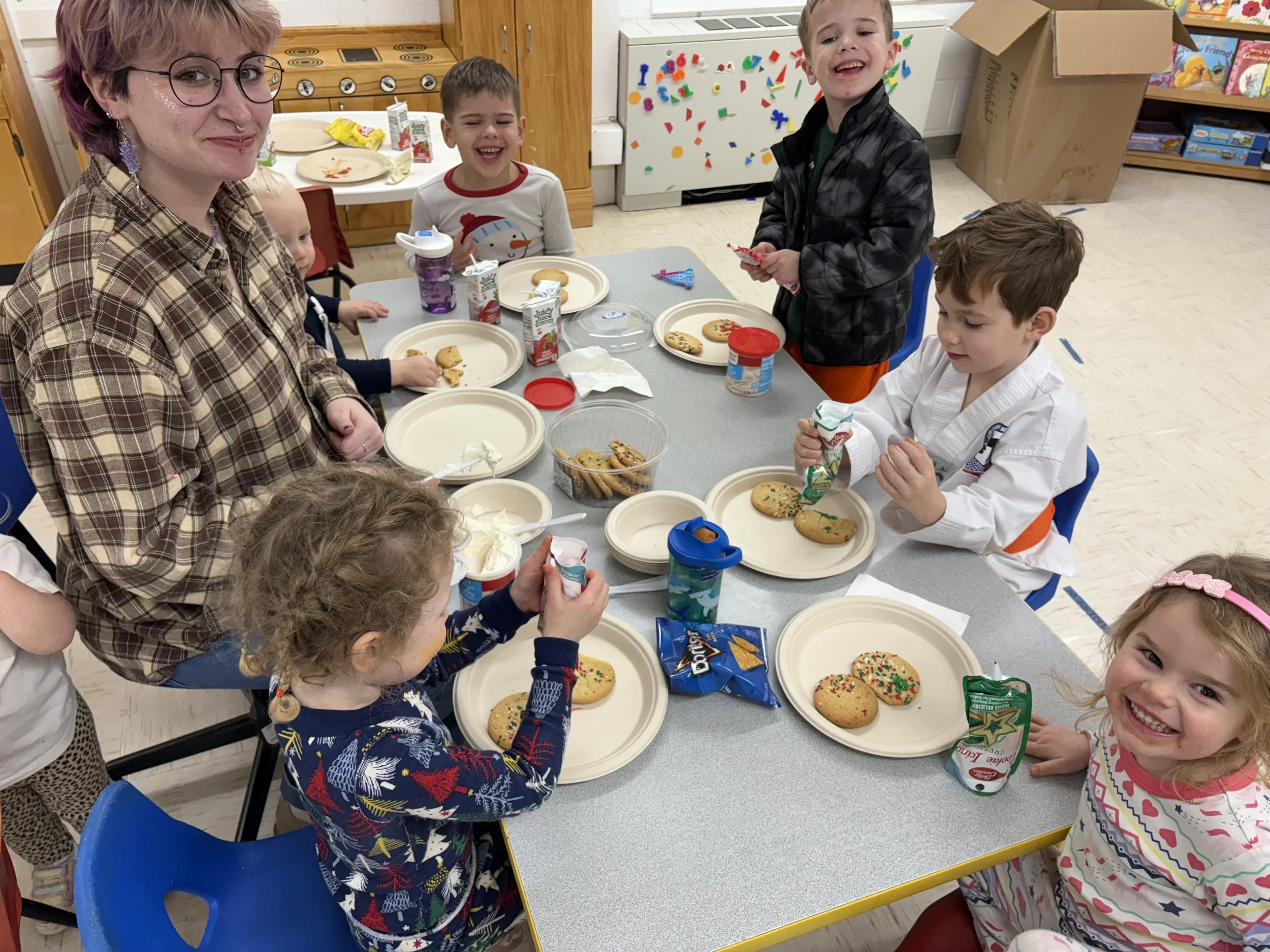 Group of children and an adult at a holiday party, sitting at a table with decorated cookies, snacks, and juice boxes, smiling and enjoying their time.