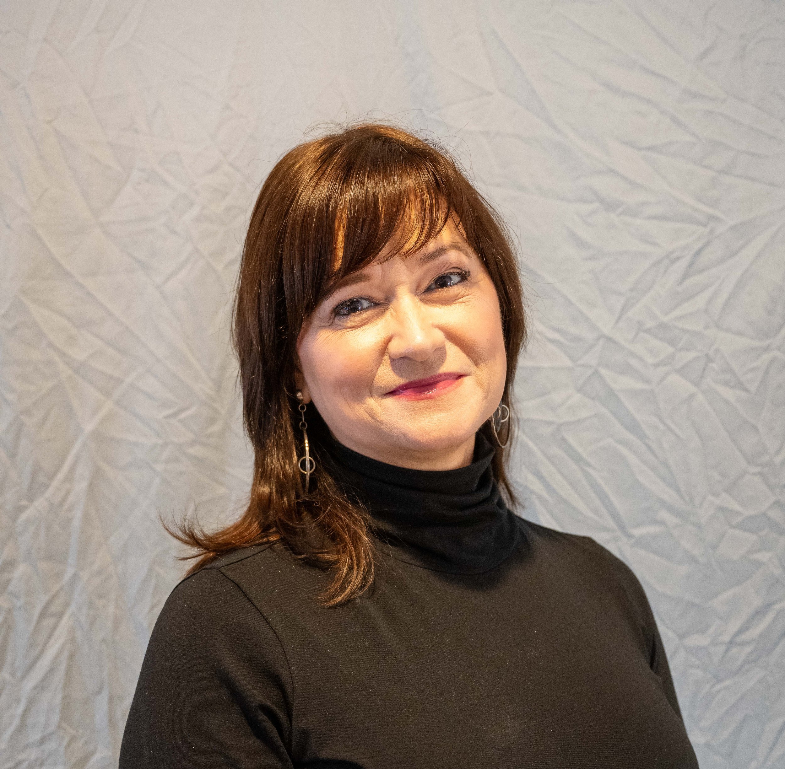 Close-up of a woman with brown hair and earrings, smiling, wearing a black top, in front of a textured white wall.