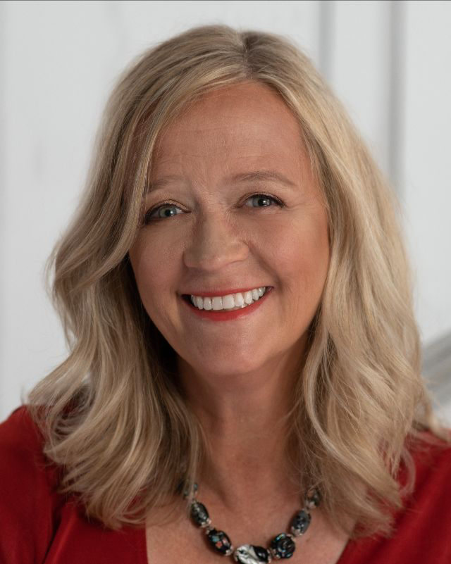 A smiling middle-aged woman with blonde, wavy hair, wearing a red top and a beaded necklace, indoors with neutral background.