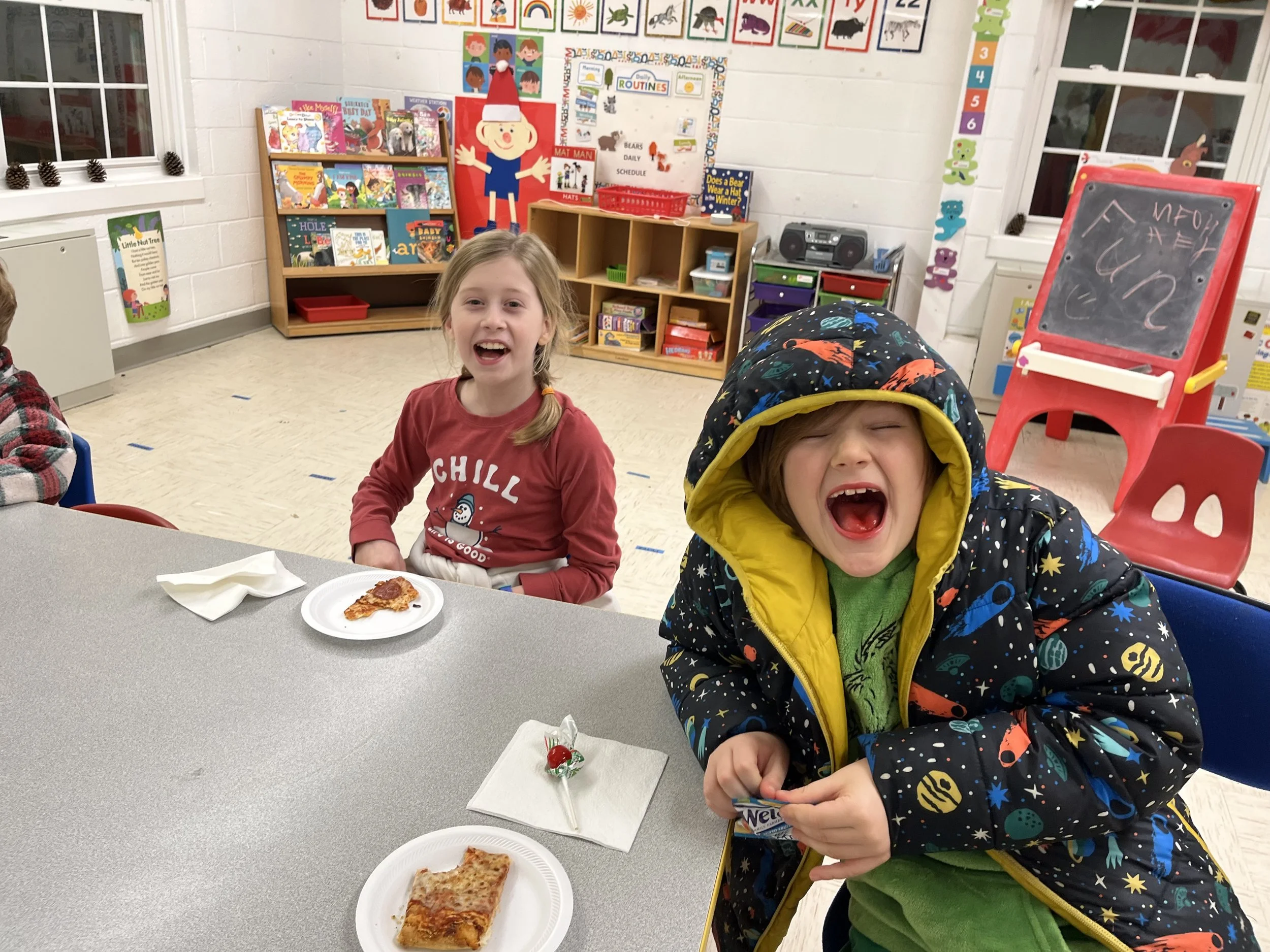 Two children at a table in a classroom, one girl with blond hair, wearing a red shirt with a snowman, and the other with brown hair wearing a colorful jacket, are smiling and laughing. Plates of pizza and a slice of pizza are on the table.
