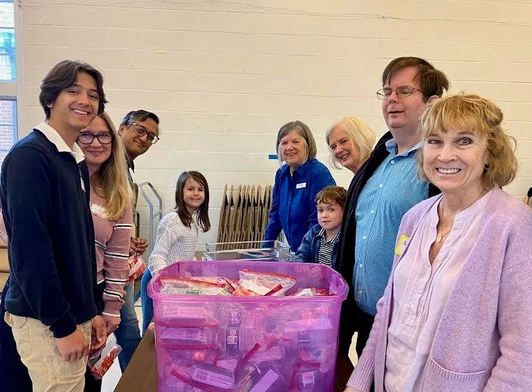 Group of smiling people, including children and adults, standing behind a table with a pink bin filled with snacks or food items in an indoor setting.