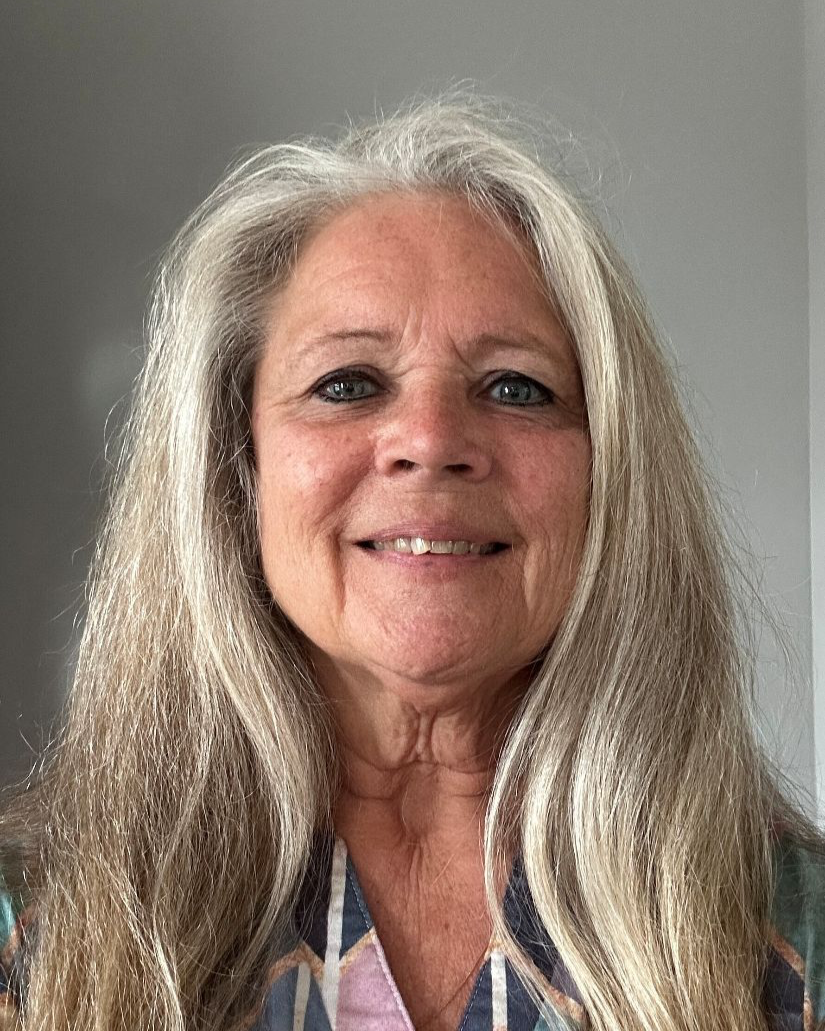 A close-up photo of an elderly woman with long gray hair, smiling, wearing a colorful striped shirt.