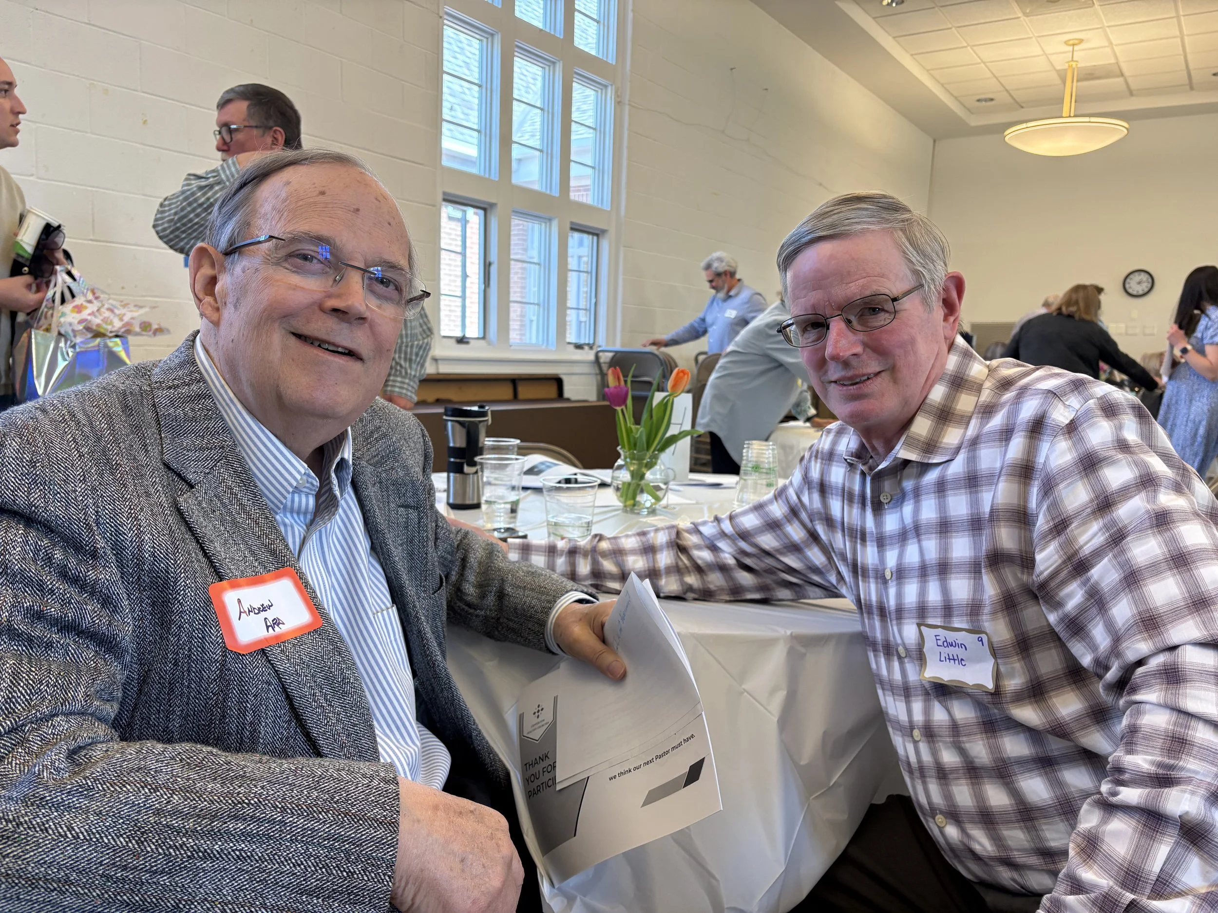 Two middle-aged men smiling at an event, seated at a table with name tags and papers. One man has a name tag saying "Aaron AR", and the other says "Edwin Little." Other people are in the background, some standing and some sitting, with a bright room and large windows.
