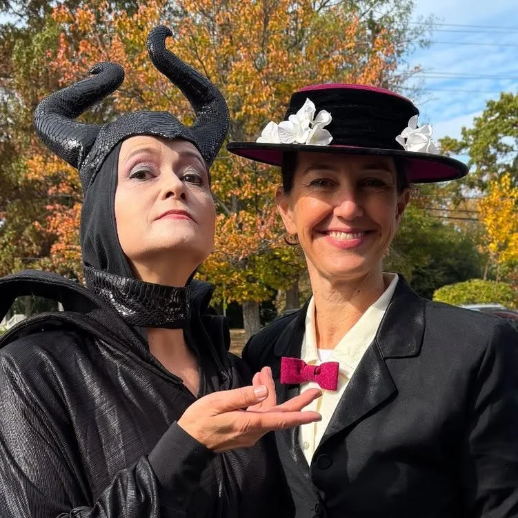 Two women in costumes posing outdoors with trees displaying fall foliage in the background. One woman is dressed as Maleficent with black horns and a black outfit, and the other woman is dressed as Mary Poppins with a black hat decorated with white flowers, a pink bow tie, and a black jacket.