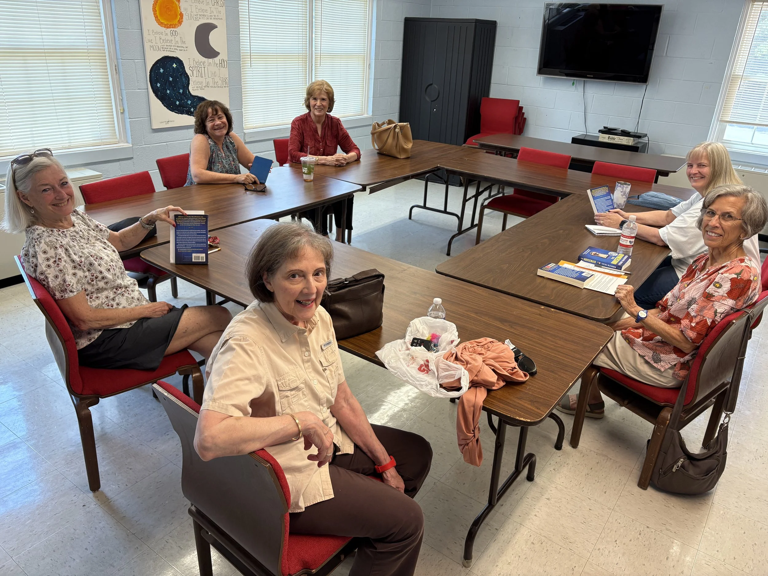 Group of seven women sitting around a large wooden conference table in a room with white brick walls, large windows with blinds, and a flat-screen TV. A colorful religious-themed poster hangs on the wall behind them.