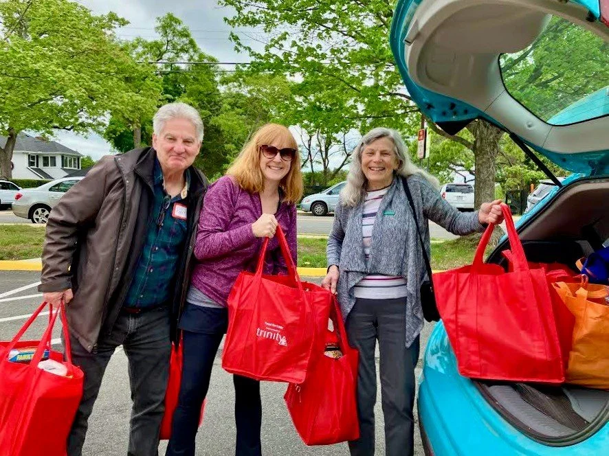 Three smiling women standing next to a blue car with an open trunk, holding red reusable bags, outdoors with green trees in the background.
