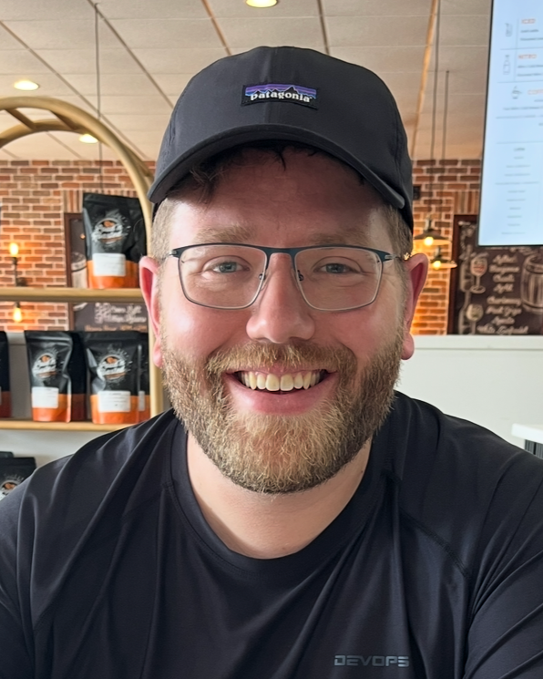 A man with glasses and a beard smiling, wearing a black Patagonia cap and a black t-shirt, sitting inside a coffee shop.