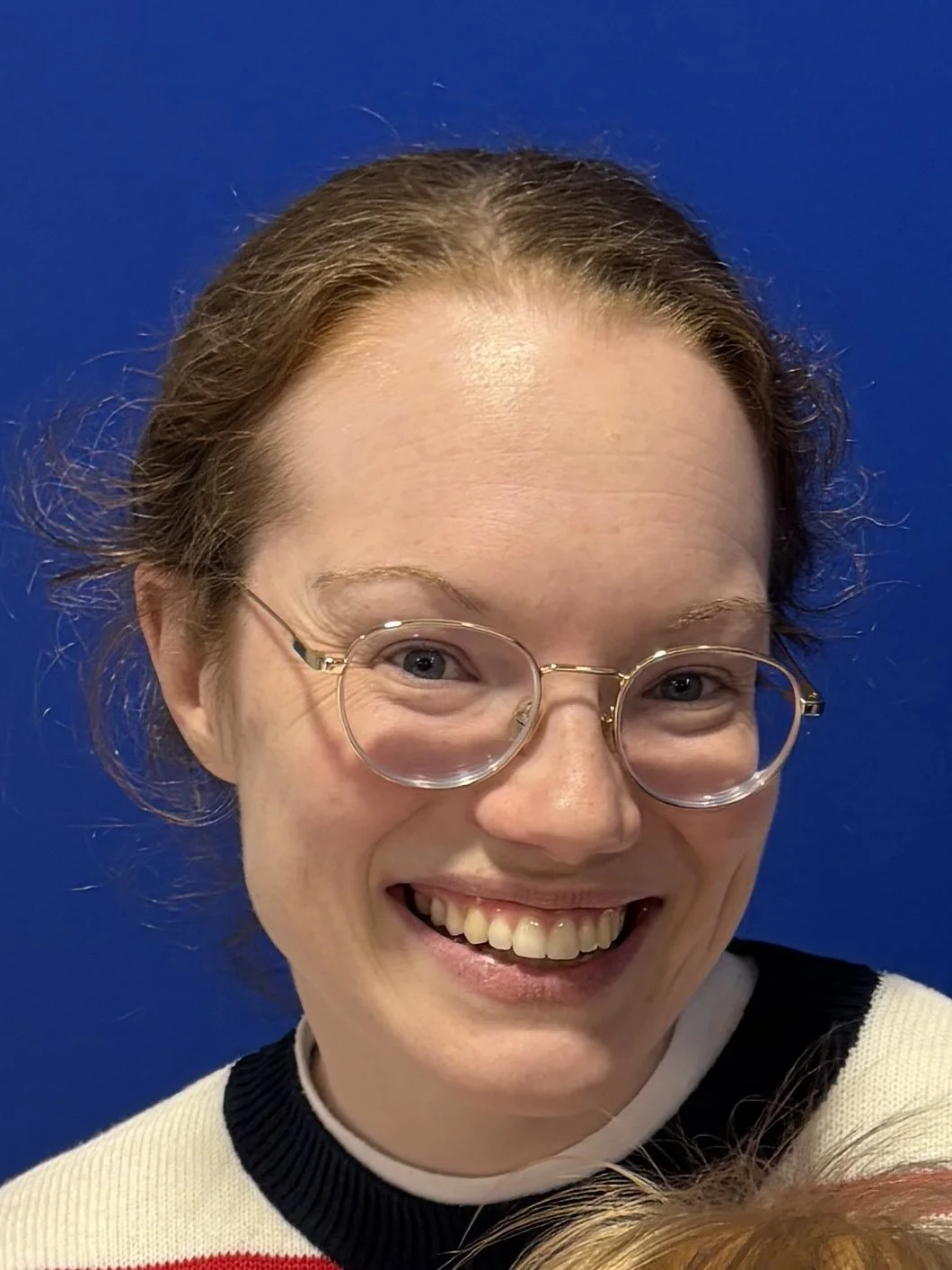 A person with short, light brown hair taking a selfie indoors. The person has piercings, tattoos, and is wearing a black shirt. The background is a textured, light-colored wall.