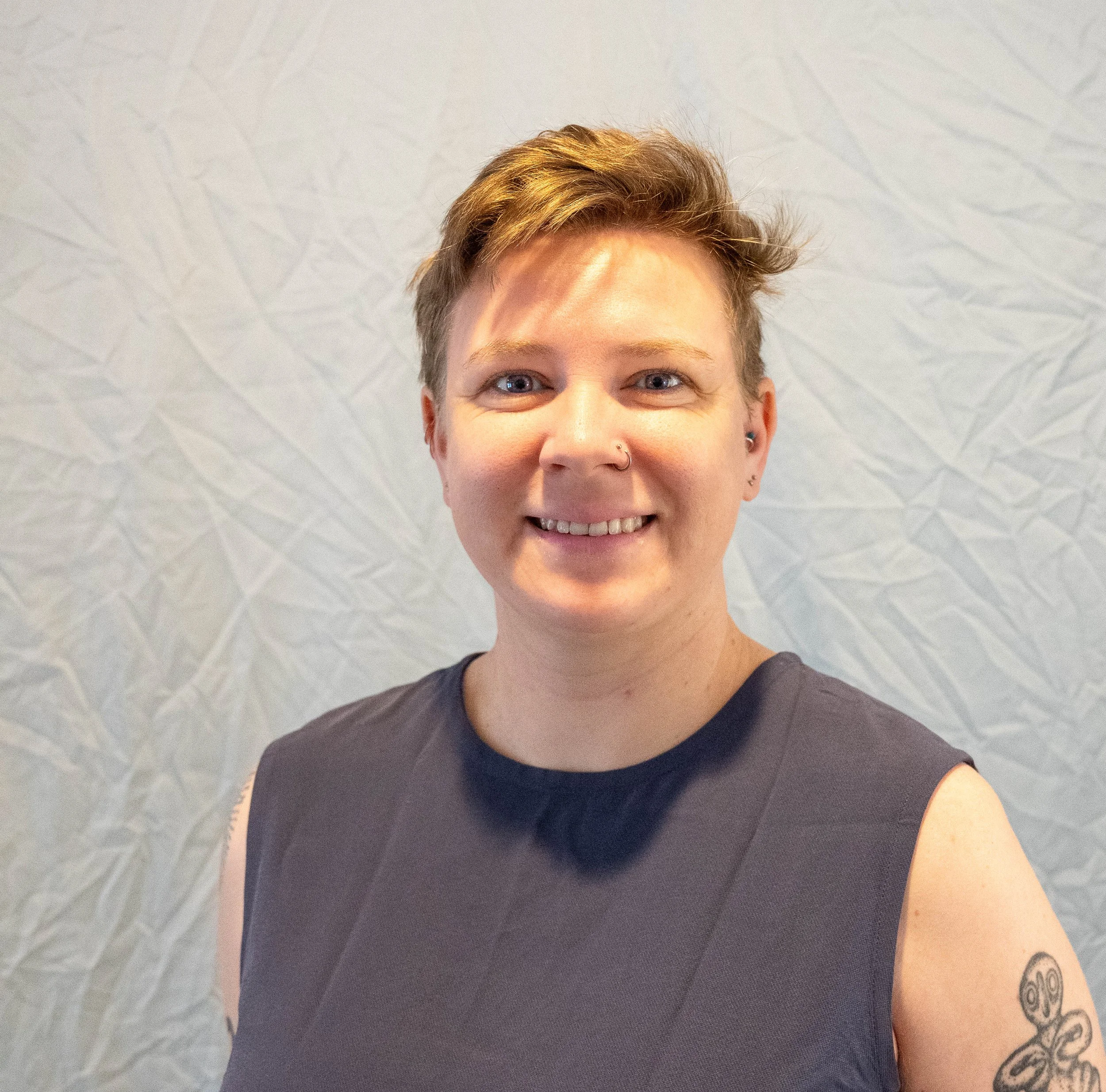 A person with short, light brown hair taking a selfie indoors. The person has piercings, tattoos, and is wearing a black shirt. The background is a textured, light-colored wall.