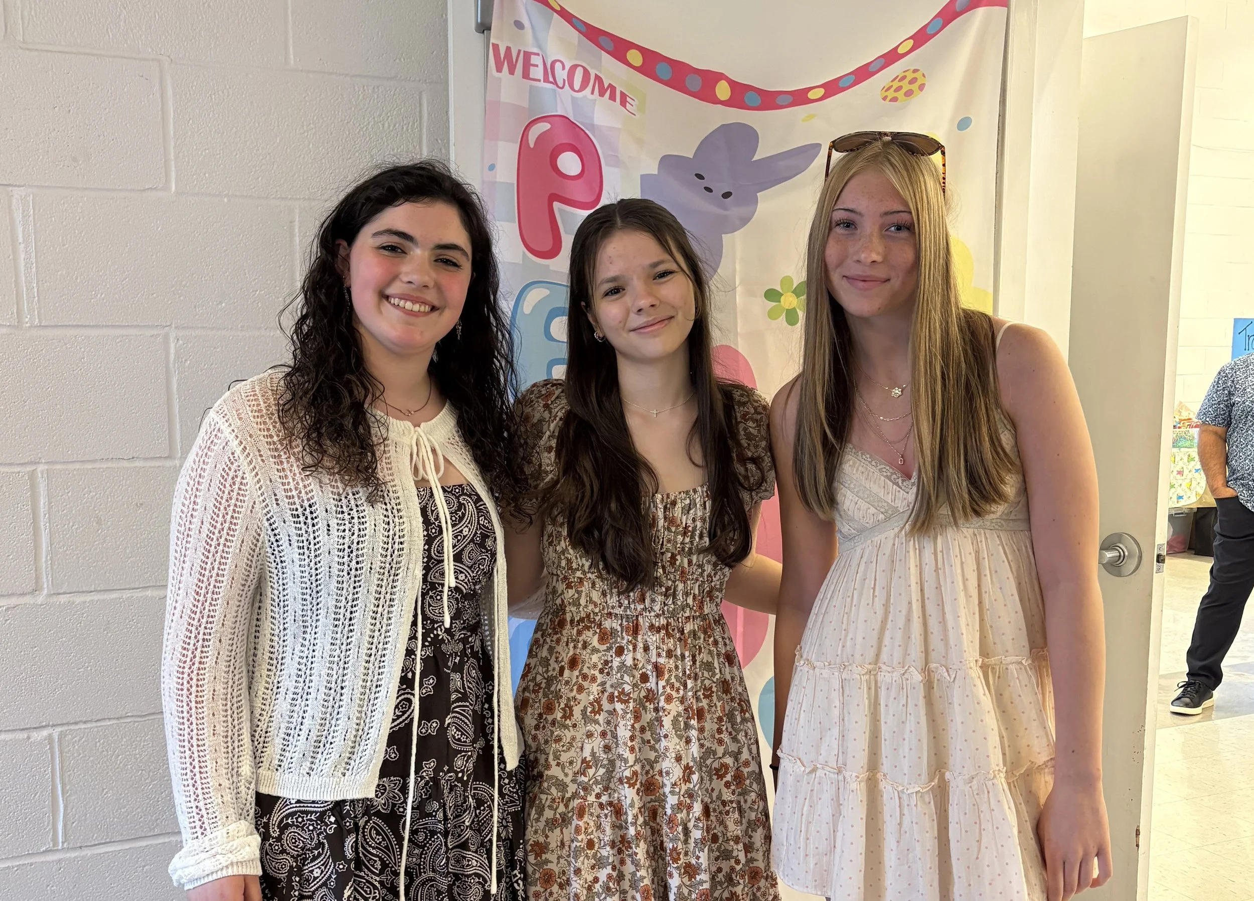 Three young women standing side by side in front of a colorful wall decoration that reads 'Welcome'. They are smiling and dressed in casual summer clothes.