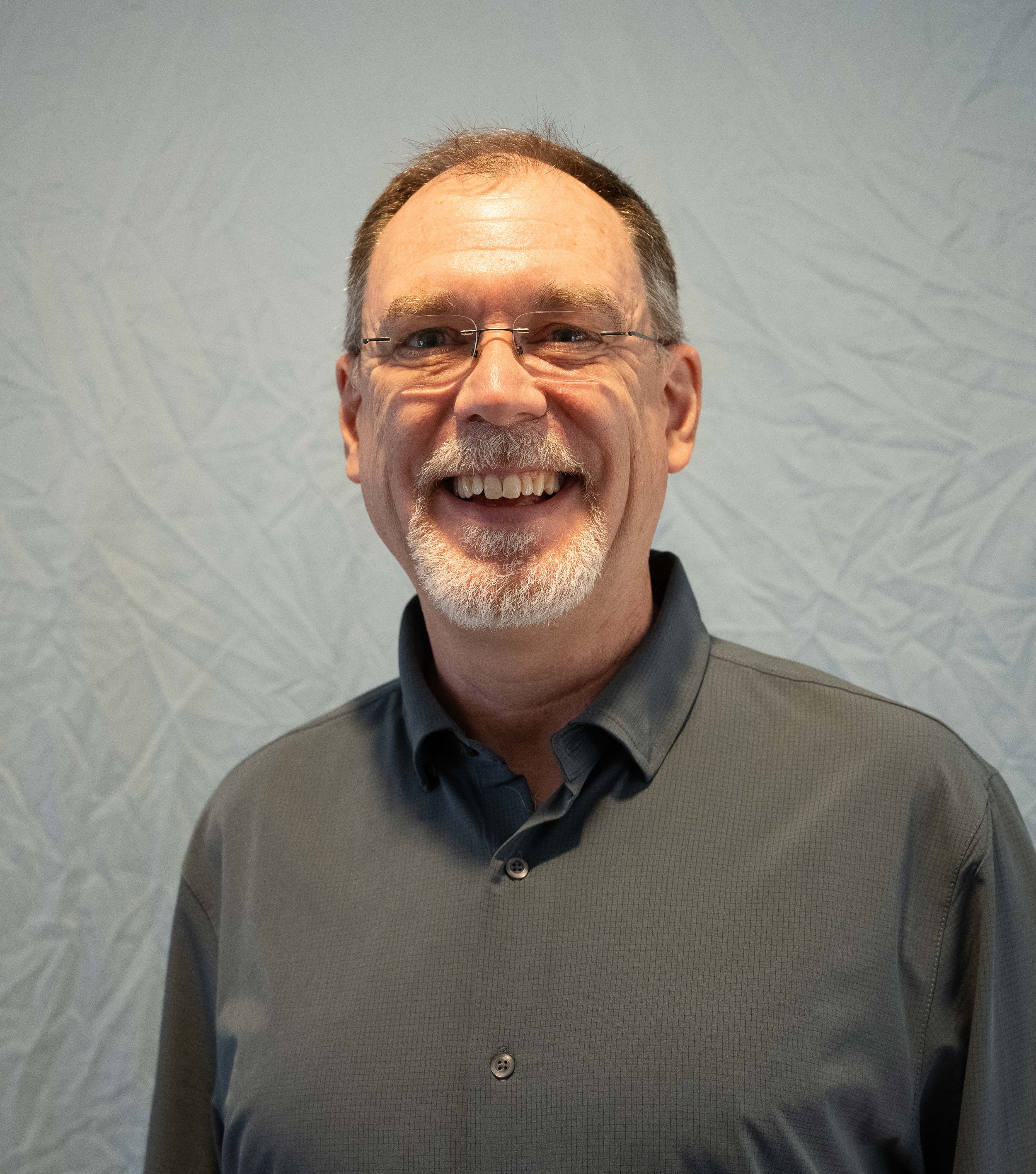 A middle-aged man with glasses, smiling broadly, dressed in a gray checkered shirt, standing indoors against a wall with subtle textured pattern.