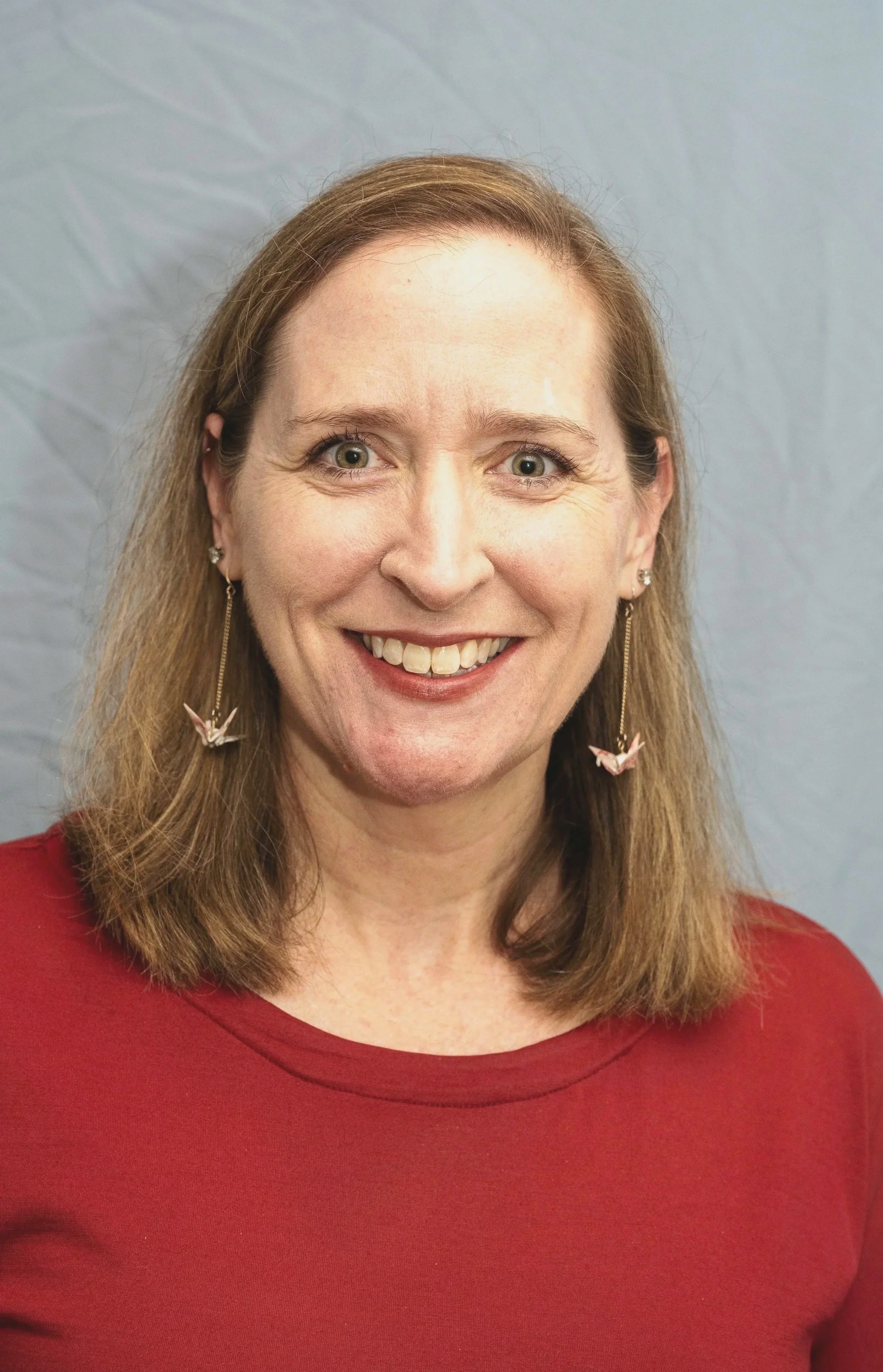 A woman with shoulder-length light brown hair and green eyes smiling, wearing earrings with origami cranes and a red top, standing in front of a plain light gray background.