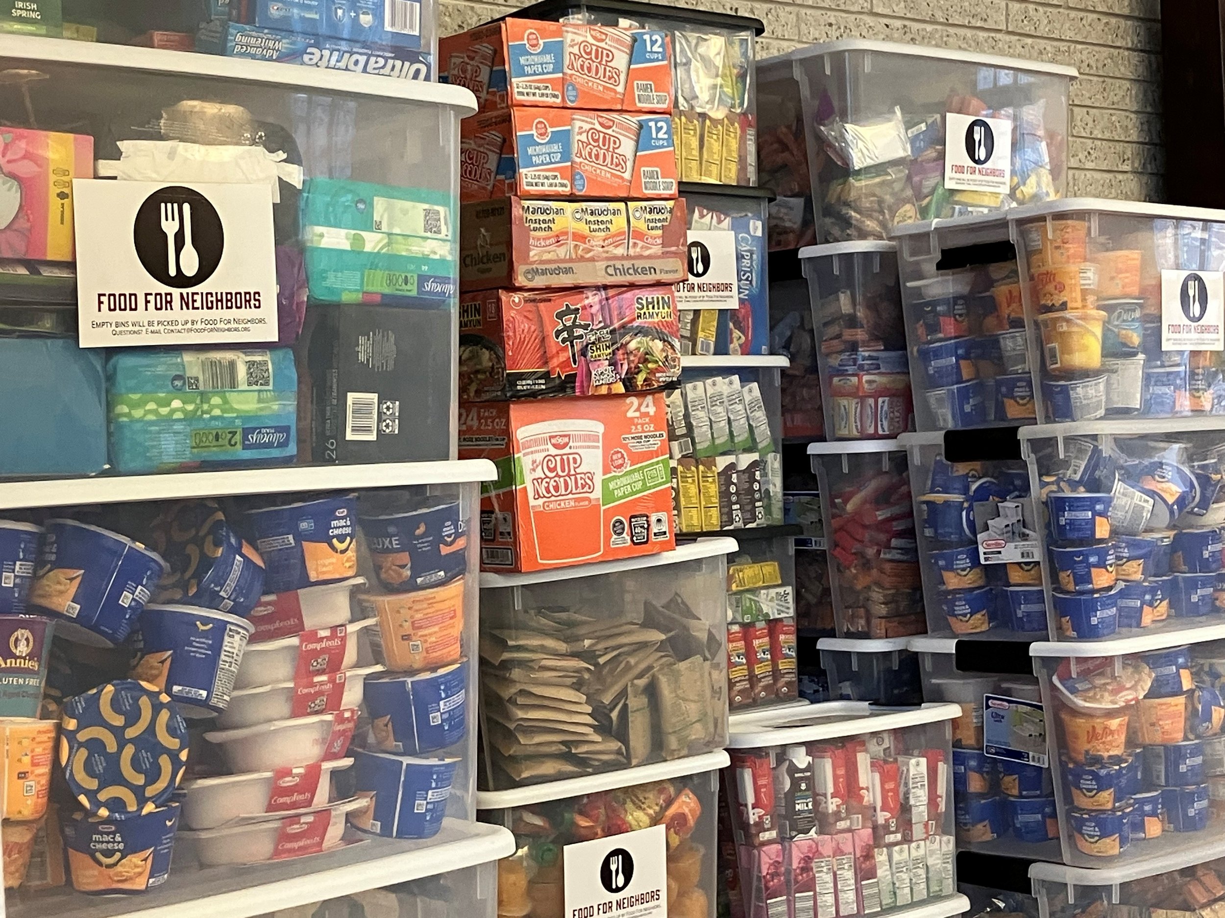 Shelves stocked with various non-perishable food items, including instant noodles, canned goods, and snack foods. There are signs indicating these food items are for neighborhood collection, with an icon of a fork and knife and the text 'Food for Neighbors'.