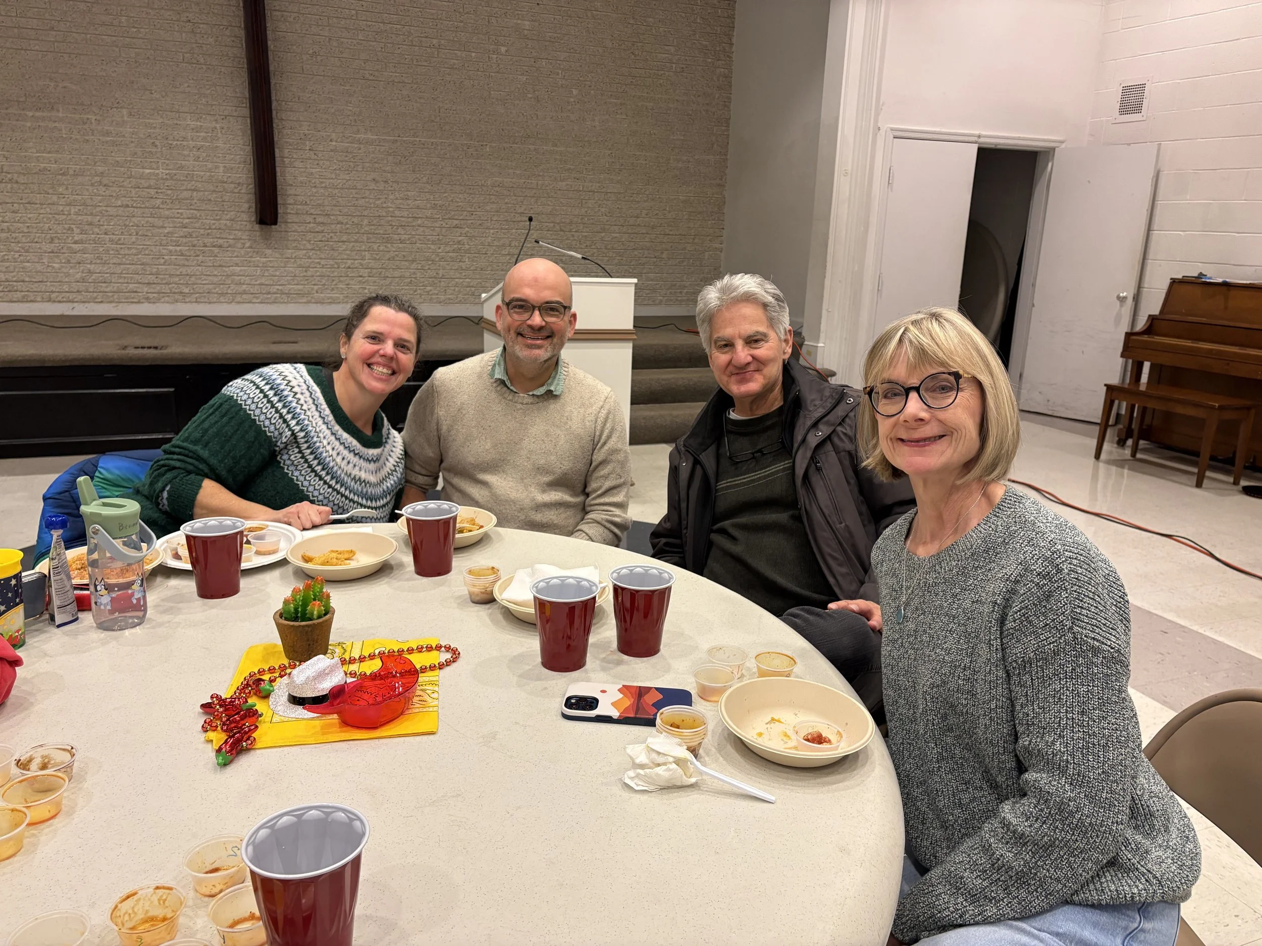 Four people sitting around a table with food, drinks, a small cactus, and holiday decoration, smiling for the camera in a large room.
