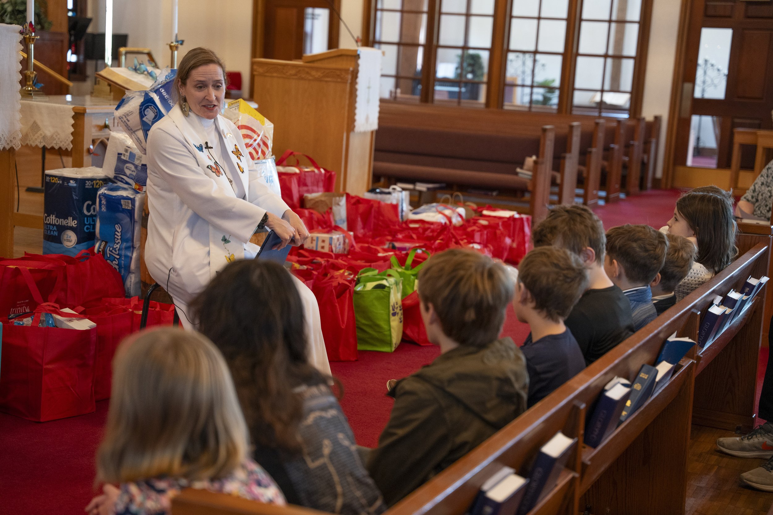 A woman in a white jacket decorated with butterfly pins is speaking to children sitting in pews inside a church. There are red and green gift bags on a table behind her, and large packs of toilet paper are also visible. The setting features wooden railings and large windows with a view outside.
