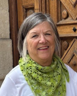 A smiling older woman with gray hair wearing a white top and a green patterned scarf, standing in front of a wooden door with decorative carvings.
