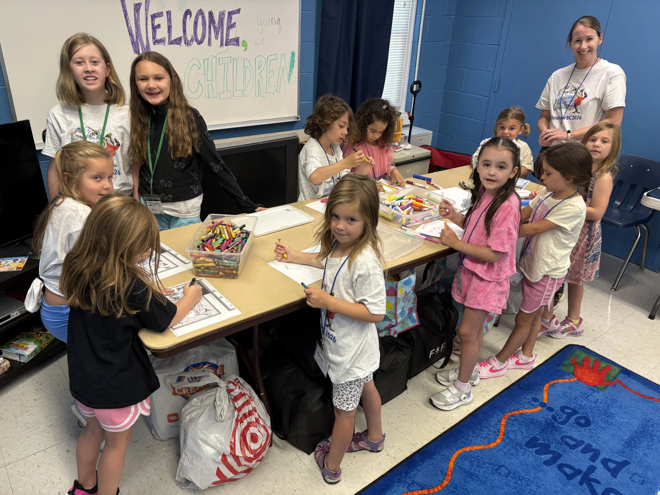 A group of young girls and a woman in a classroom, gathered around a table with markers and coloring sheets. There is a large welcome sign on the whiteboard behind them that reads 'Welcome going ice children.' The girls are smiling and appear to be engaged in a craft activity.