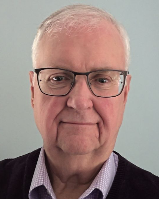 A close-up of an older man with white hair, glasses, wearing a collared shirt and a dark sweater, smiling slightly against a plain background.