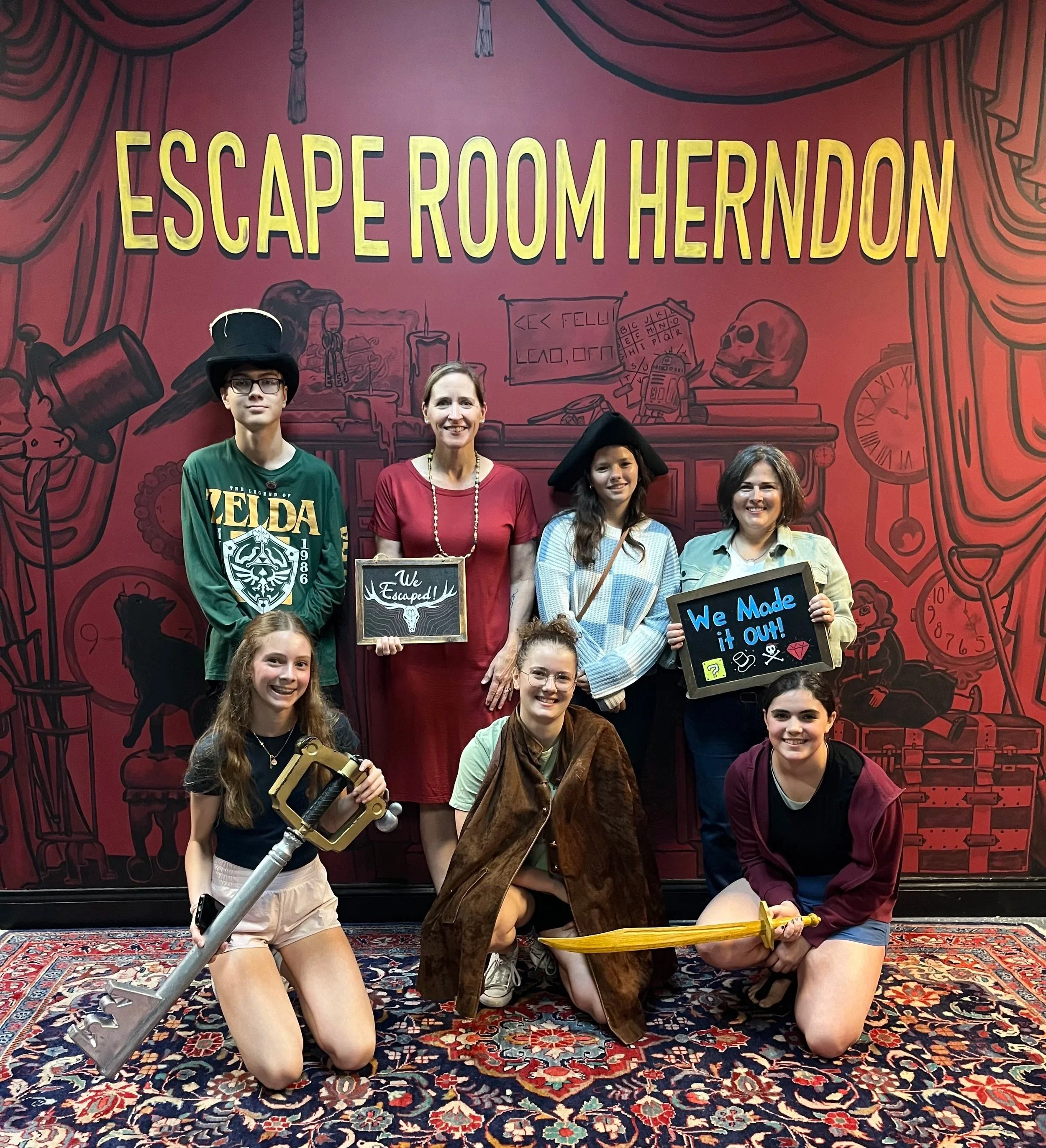 A group of people dressed in costumes, standing in front of a red backdrop at Escape Room Herndon. Two women are holding signs that say "We Escaped!" and "We Made it Out!".