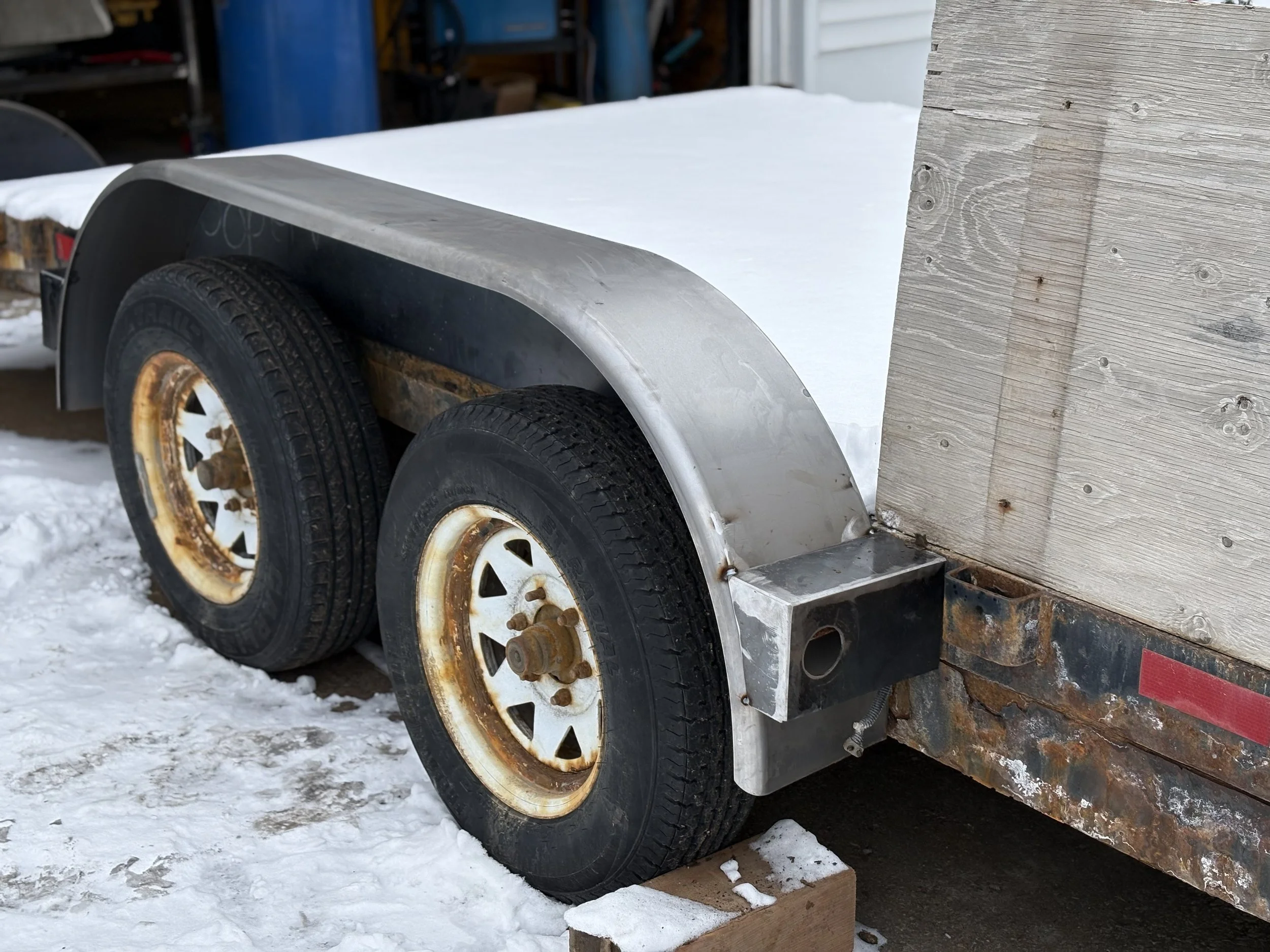 A close-up of a utility trailer with black tires and rusted white rims, parked on snow in front of a wooden structure with visible screws.