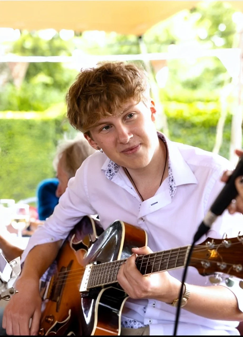Young man with light brown hair playing an acoustic guitar at an outdoor event, smiling and looking at the camera, with a blurred green garden background.