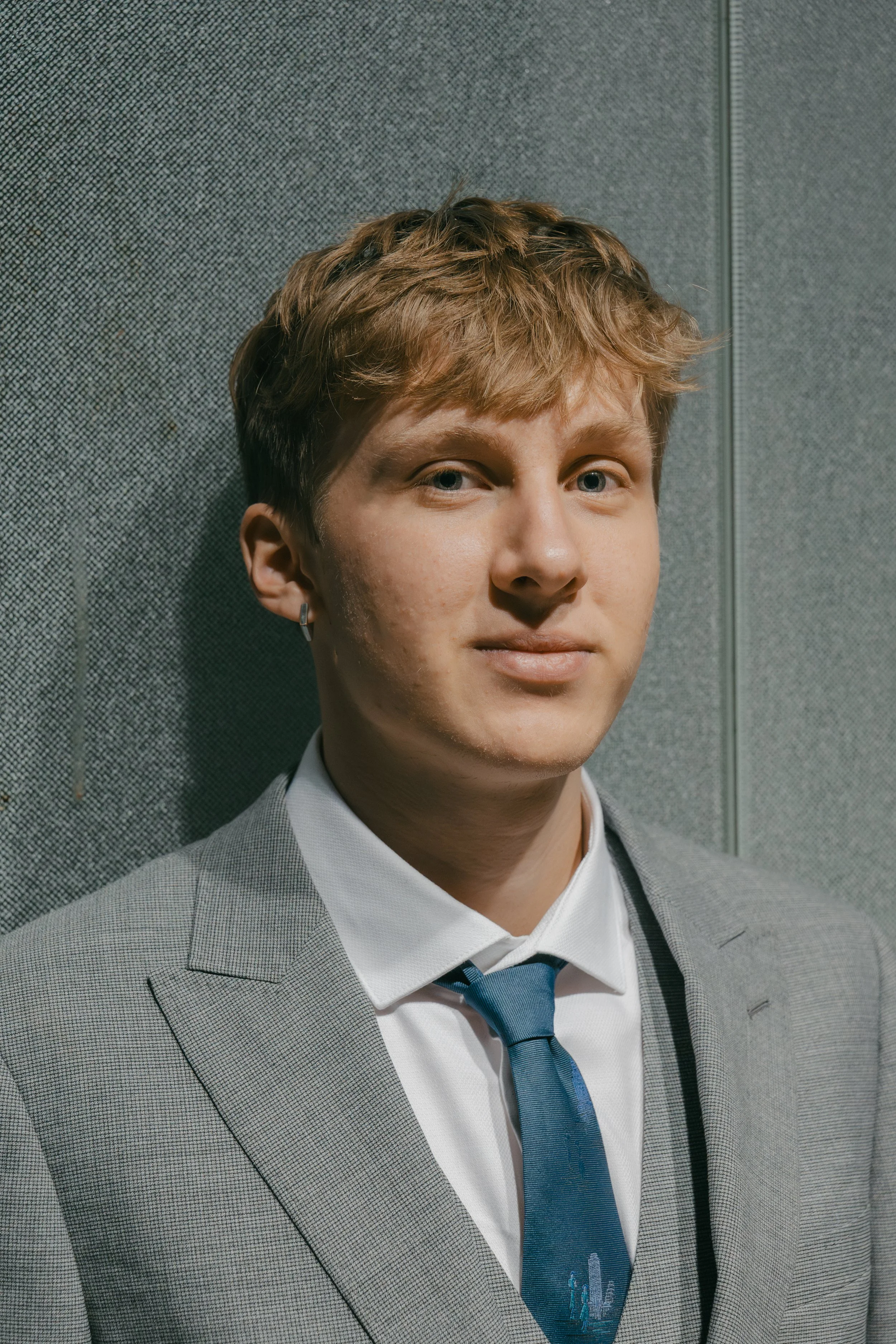 A young man with light hair, wearing a gray checkered suit, white shirt, and blue tie, standing against a gray textured wall.