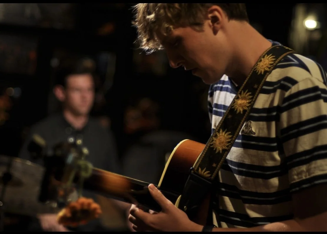A young man playing an acoustic guitar in a dimly lit room, with a guitar strap featuring sunflowers, and a blurred background of another person.