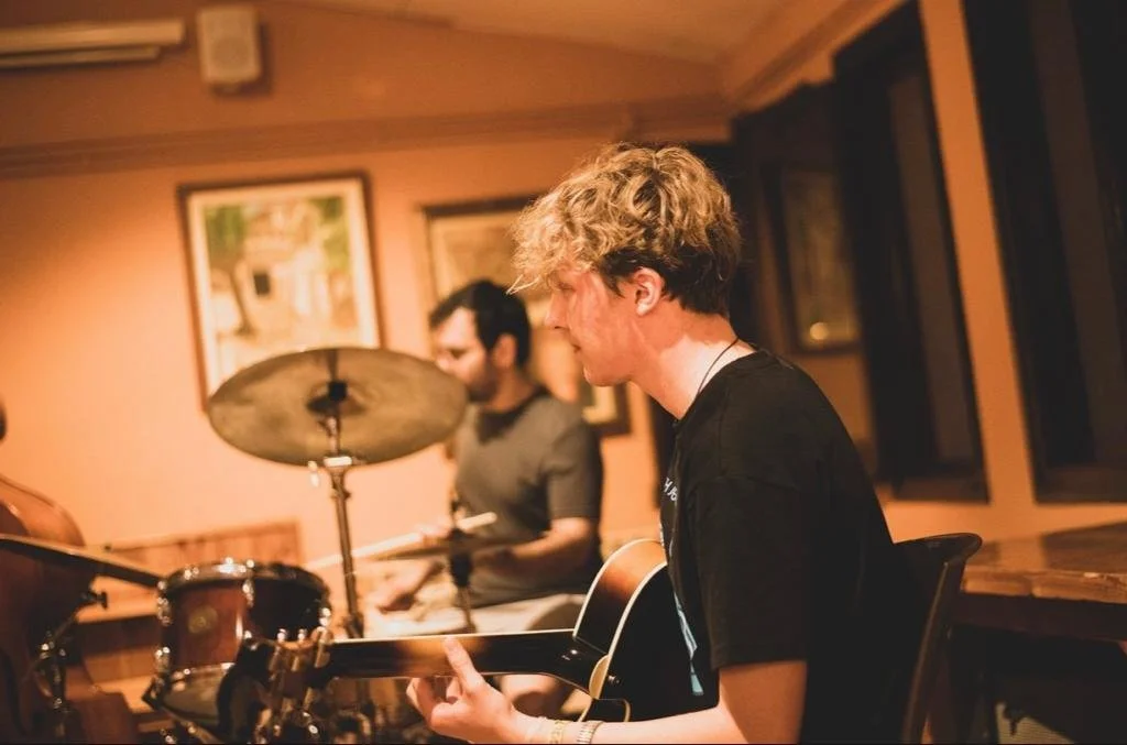 Two young men playing musical instruments in a cozy room with warm lighting, one on guitar and the other on drums.
