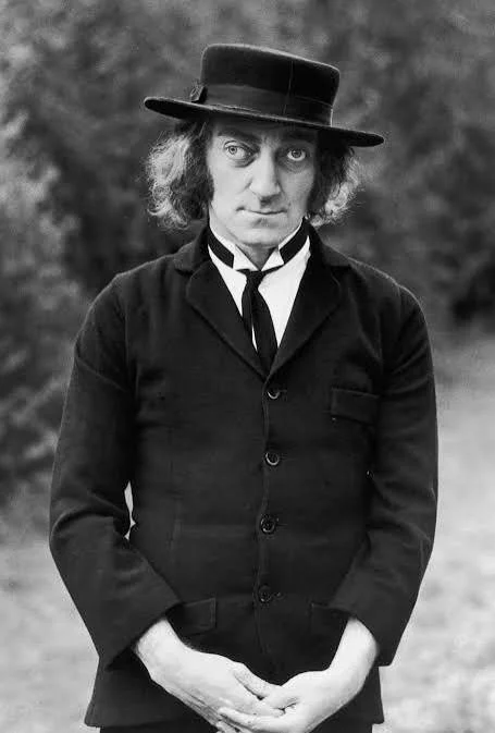 A black and white photo of a man in vintage clothing, wearing a dark suit, a white shirt, a necktie, and a wide-brimmed hat. The background appears to be an outdoor natural setting.