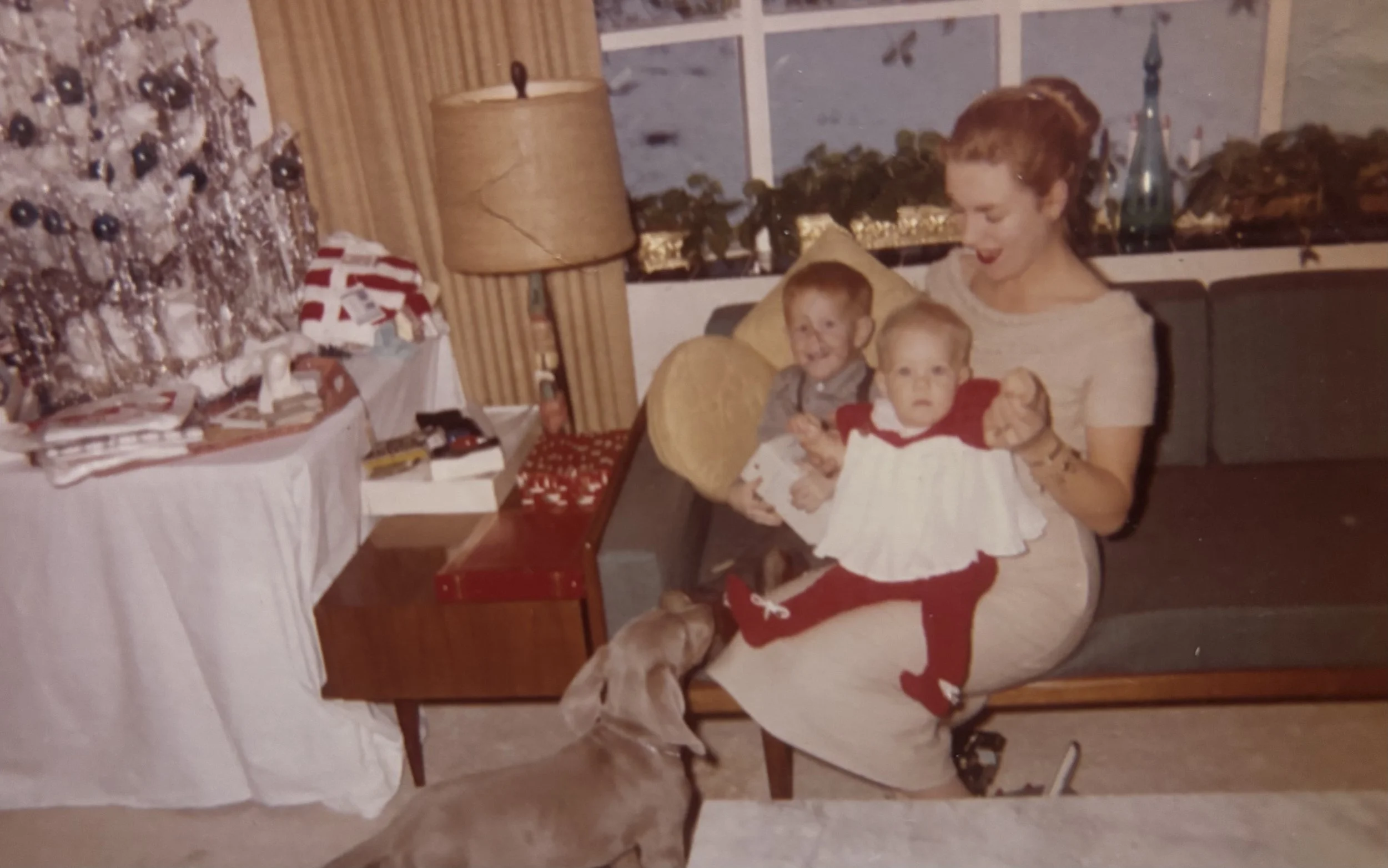A woman with two young children sitting on a couch during Christmas, with a decorated Christmas tree and wrapped presents on a table nearby.