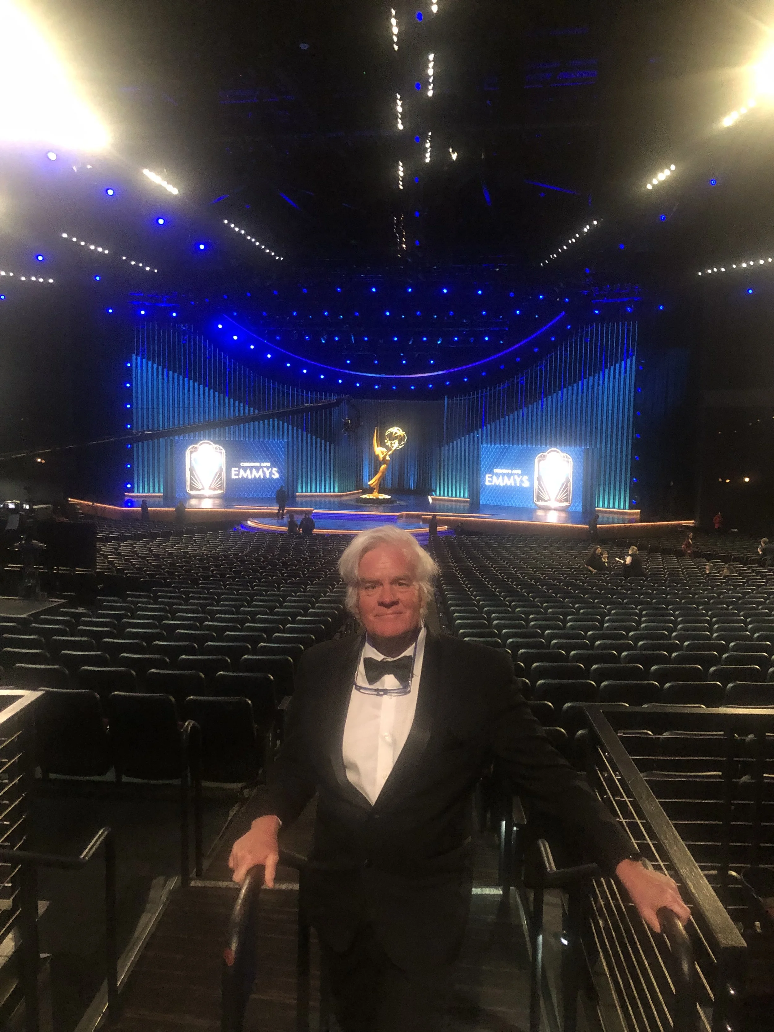 A man in a tuxedo holding a guitar in an empty theater or awards ceremony hall with a stage, blue lighting, and large screens displaying "EMMYS".