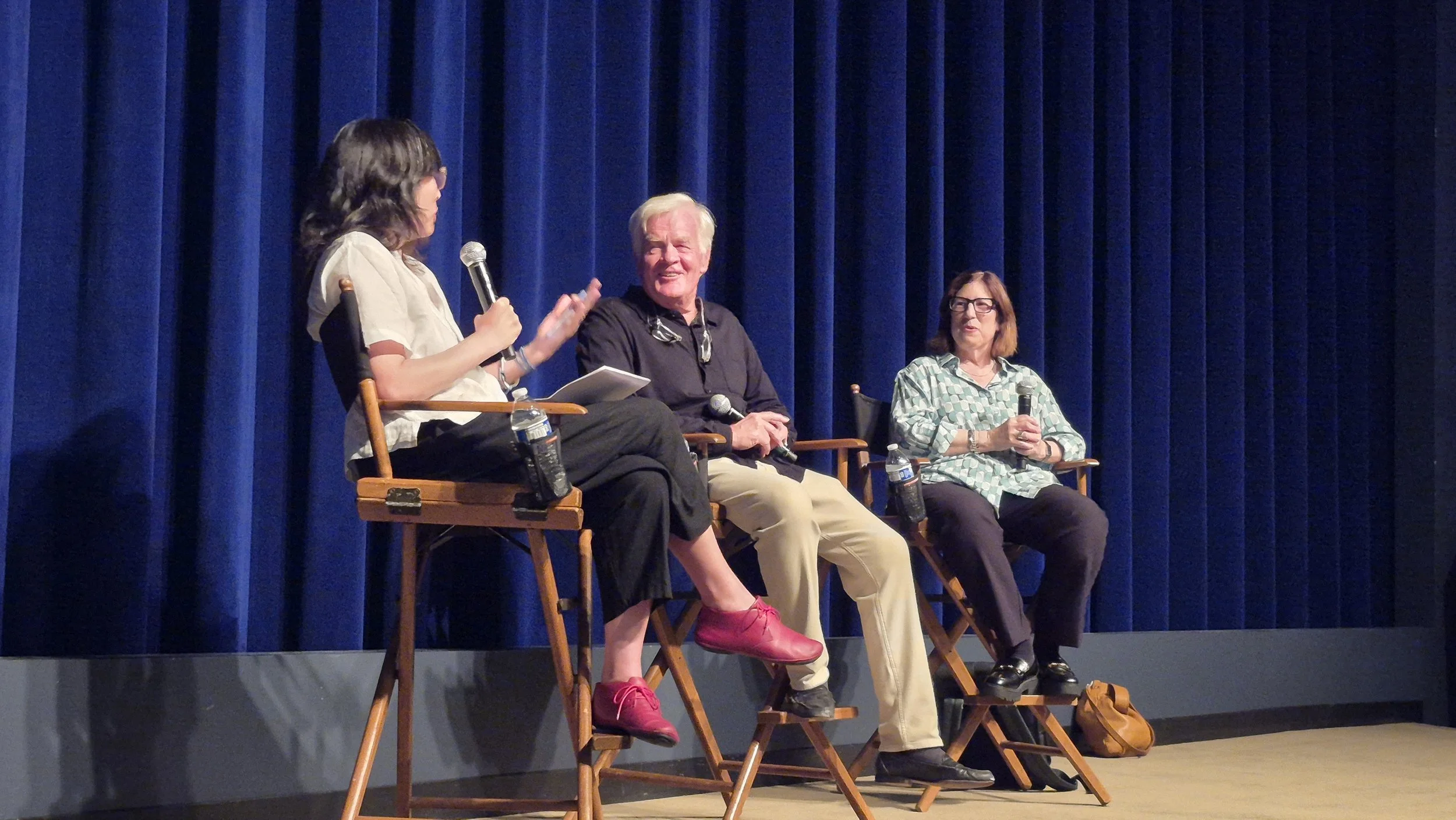 Three people seated on a stage engaging in a panel discussion. The woman on the left is speaking into a microphone and gesturing with her hand. The man in the middle is smiling and holding a microphone. The woman on the right is holding a microphone 