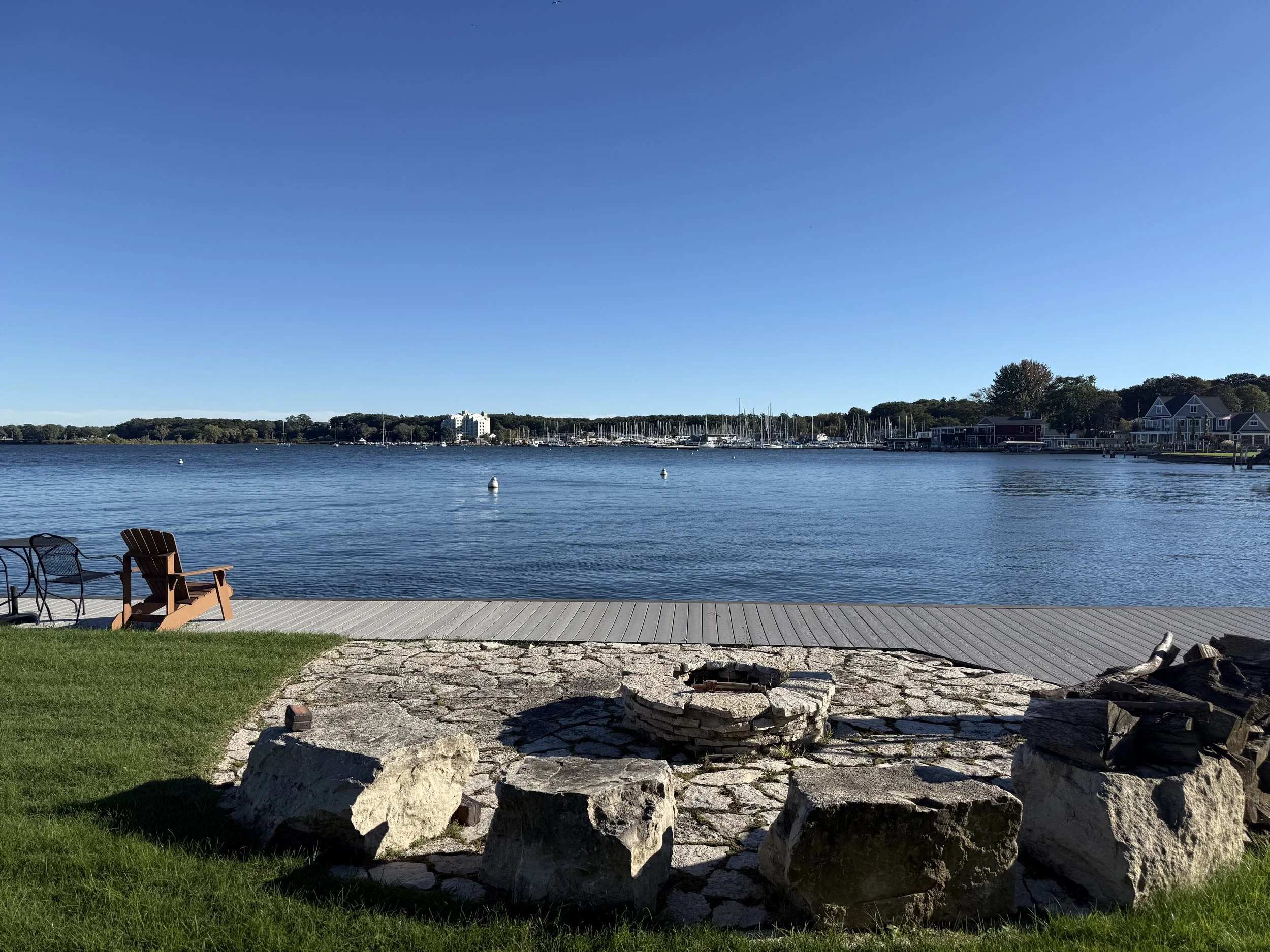 A lakeside scene with a wooden dock and chairs on the grass, calm water with boats, trees, and buildings in the background under a clear blue sky.