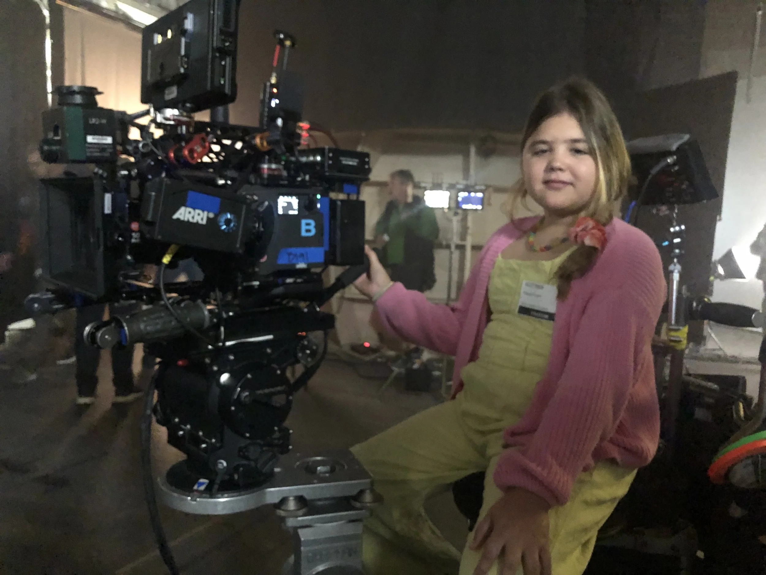 A young girl in a pink jacket and yellow dress operating a camera on a film set or studio, with multiple monitors and people in the background.
