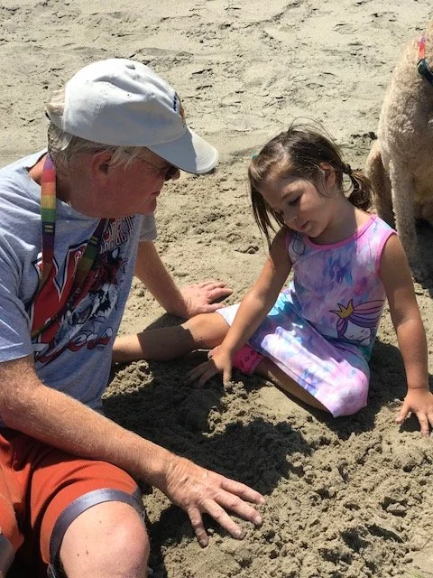 An elderly man and a young girl sitting on the sand at the beach, playing together with a small mound of sand.