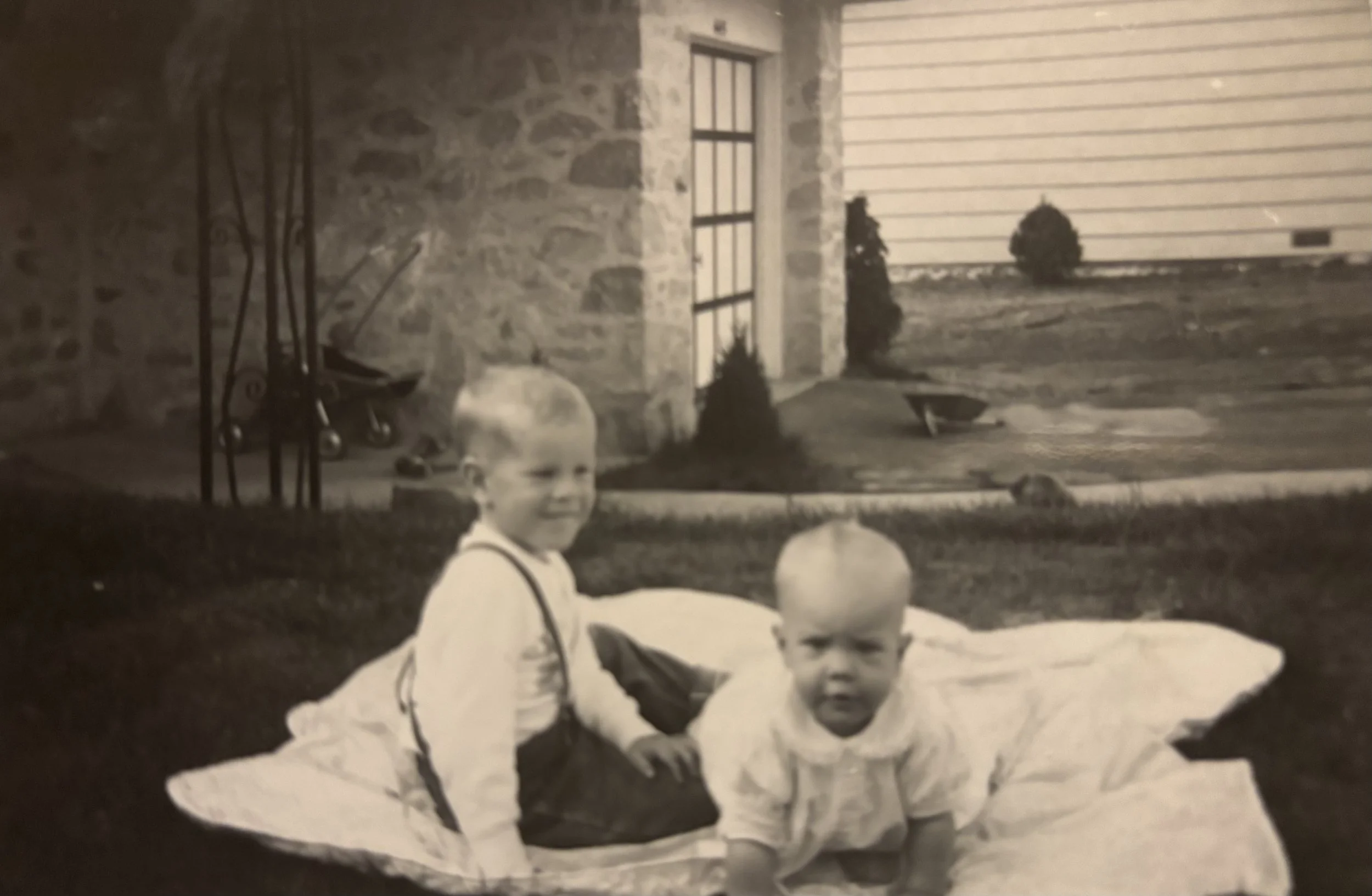 Black and white photo of two young boys with bald heads sitting at a table outdoors, smiling, with balloons nearby. In the background, there is a stone wall, a window with bars, a garage door, and some potted plants or bushes.