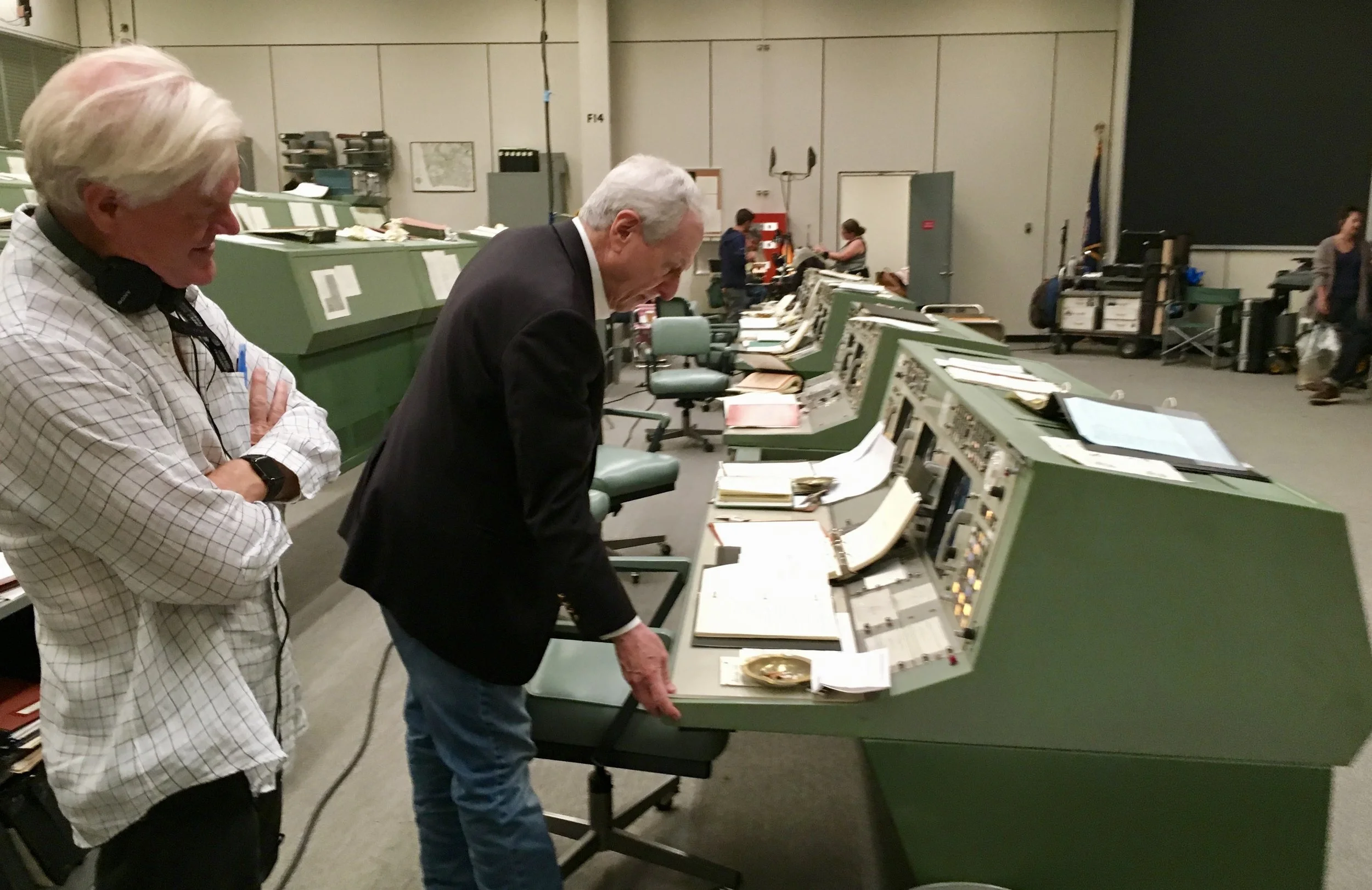 Two men and a woman look at a control panel in a room with several other people and equipment.