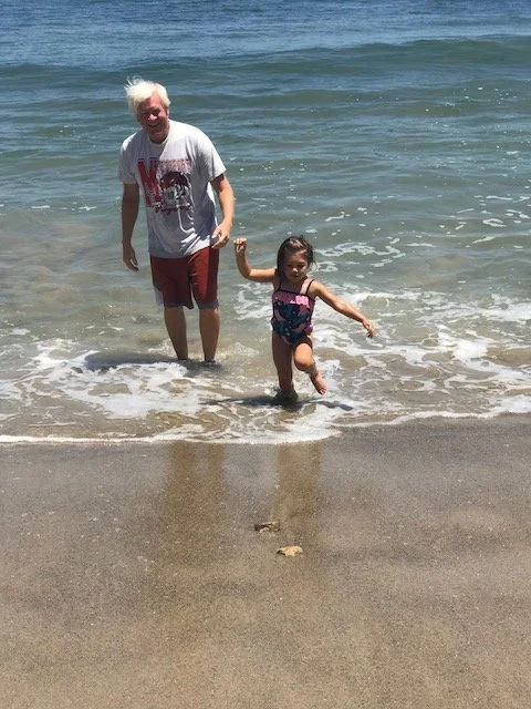 An elderly man and young girl playing and holding hands at the beach near the water.
