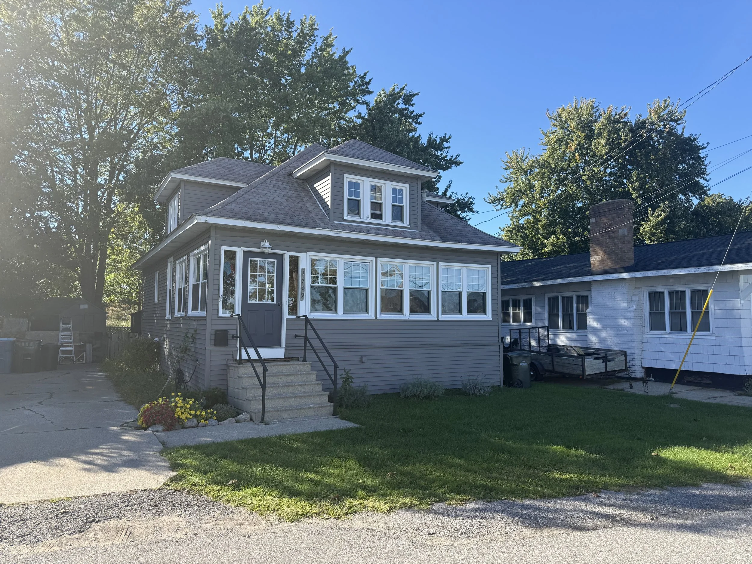 Front view of a house with gray siding, multiple windows, a gabled roof, and a small porch with stairs. There are flowers in a small garden in front of the house, and a neighboring house with a brick chimney is partially visible. Tall trees and a cle