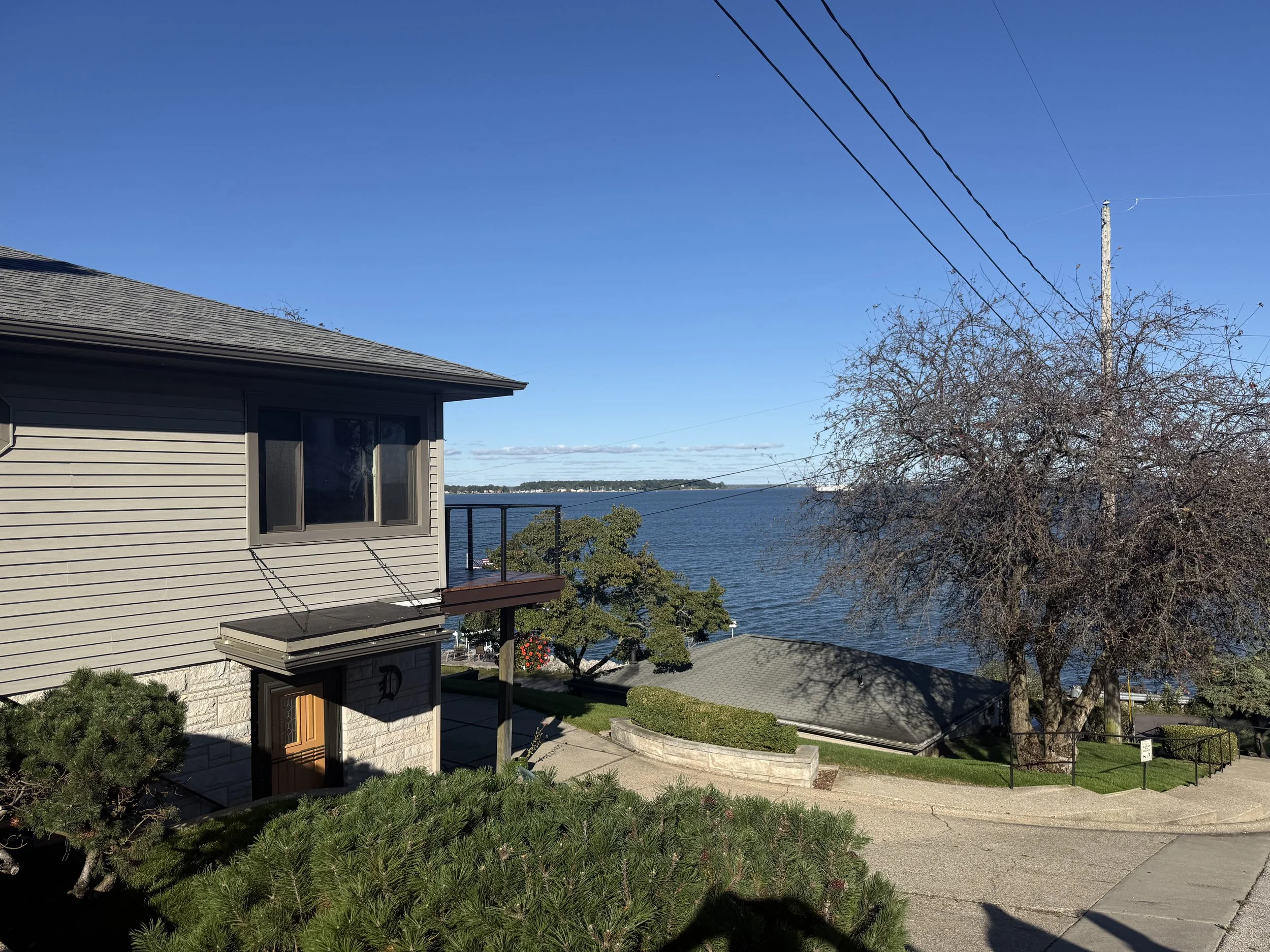 View of a house with beige siding and a brown door, overlooking a body of water with a bridge in the distance, under a clear blue sky.