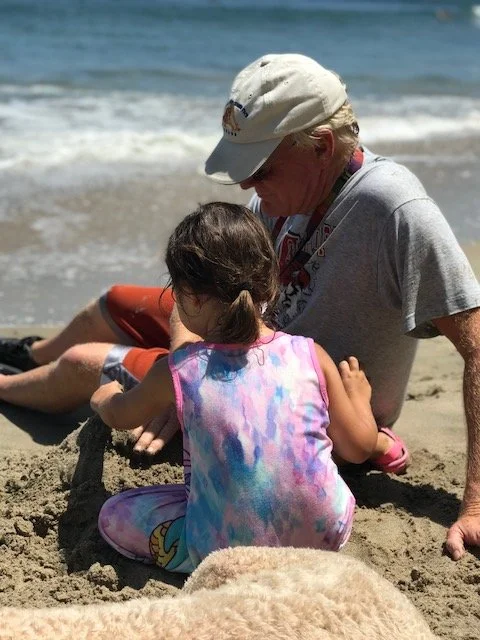 A man and a young girl sitting on the sandy beach near the water, with the man looking down at the girl who is playing in the sand.