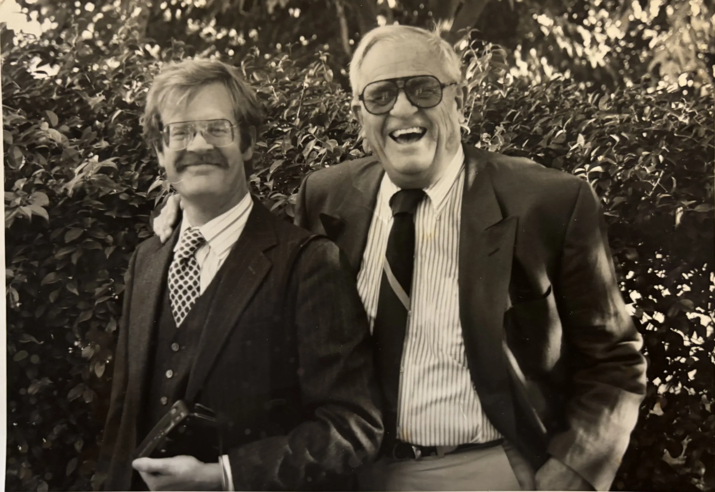 Two older men in suits and glasses smiling and laughing outdoors, surrounded by leafy bushes.
