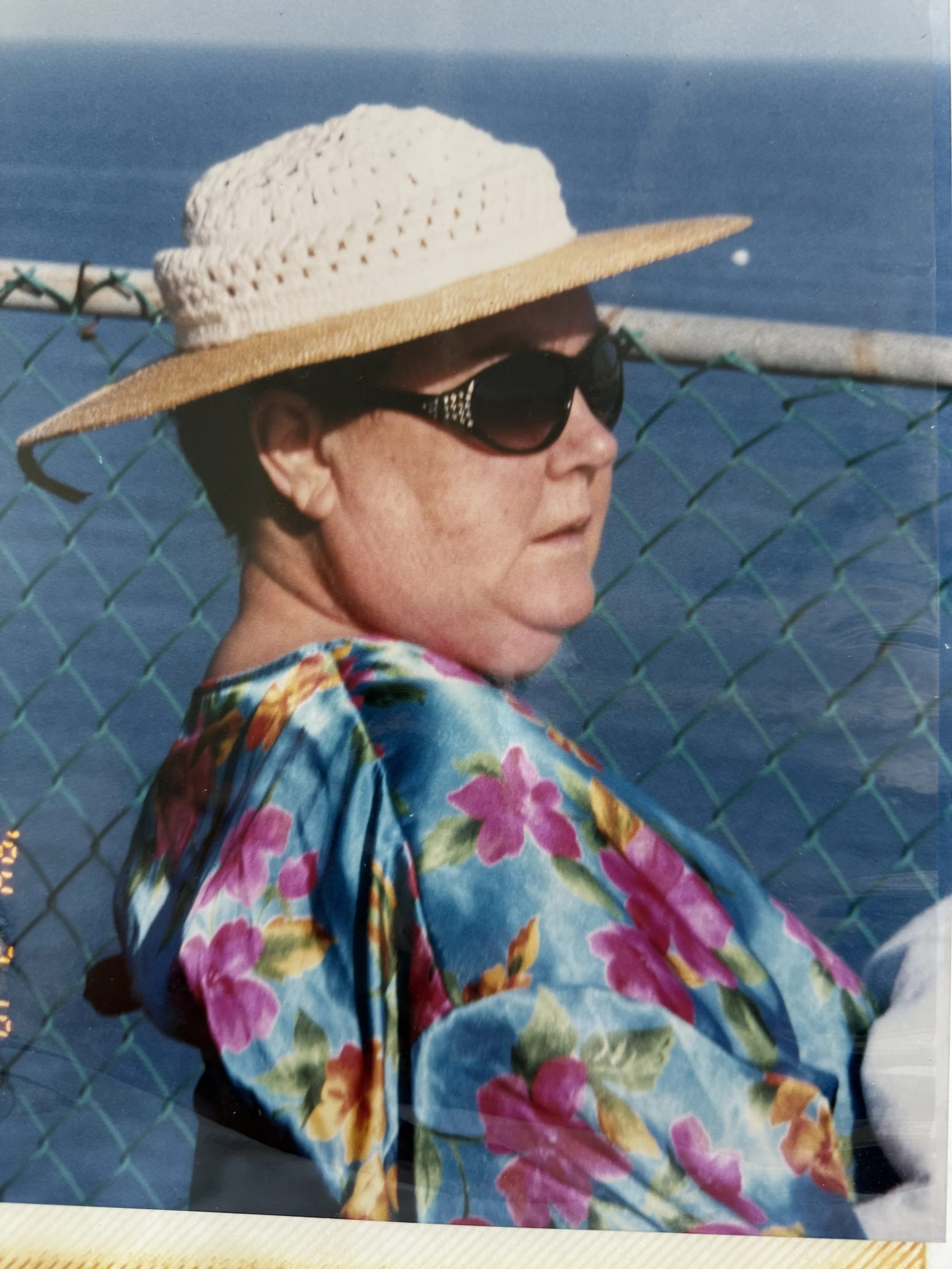 A person wearing large black sunglasses, a wide-brimmed straw hat with a crochet top, and a vibrant floral shirt standing in front of a blue ocean with a chain-link fence.
