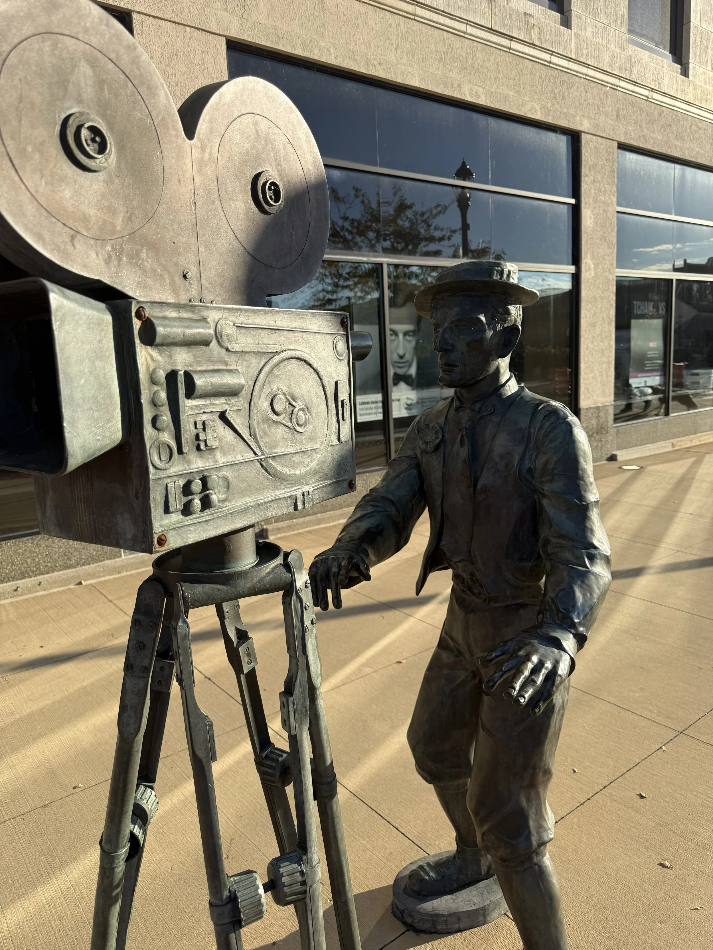 A bronze statue of a man in vintage clothing operating an old-fashioned movie camera on a tripod, placed outdoors in front of a building with large windows reflecting the sky and trees.