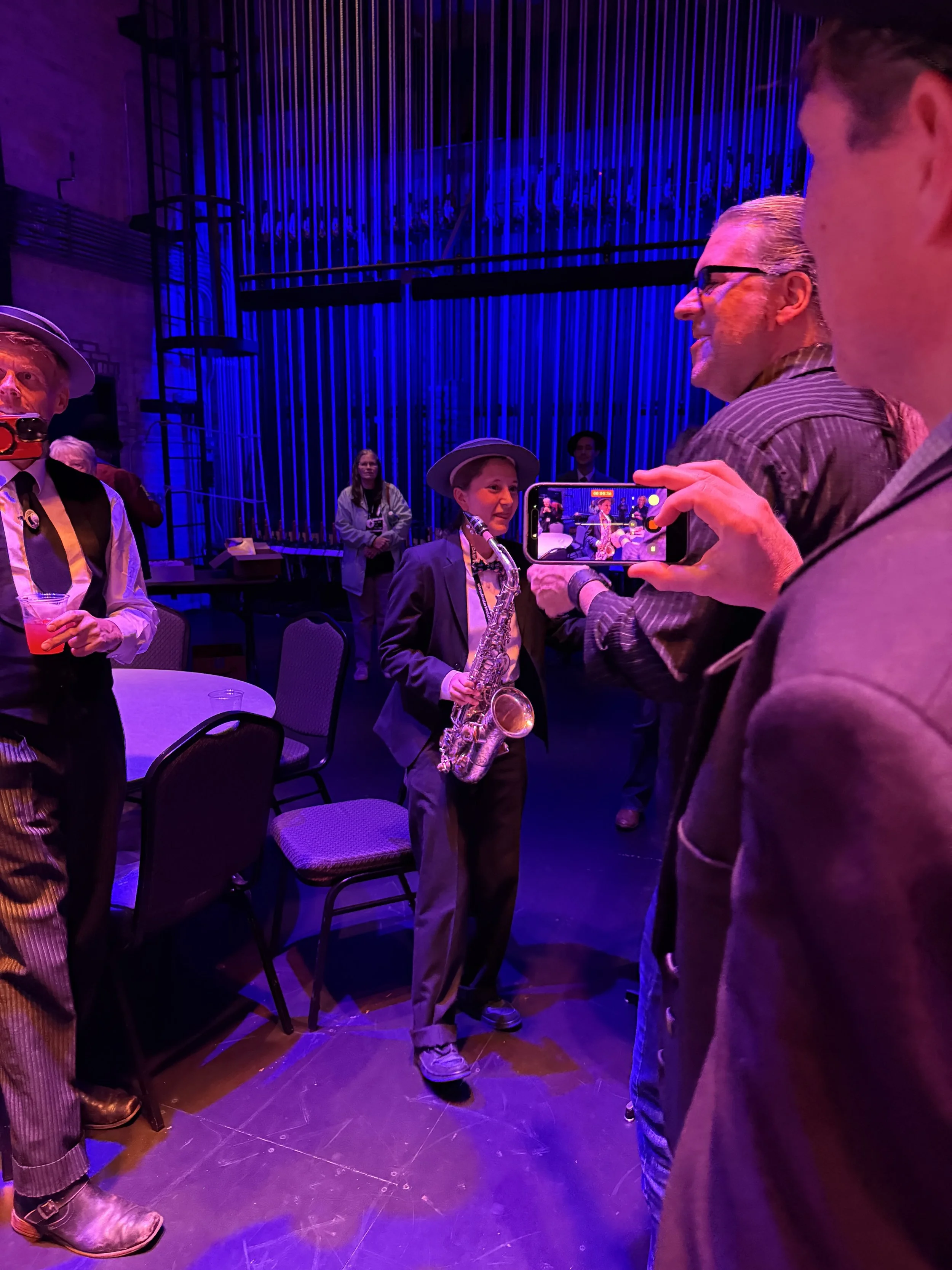 Group of people standing and talking in a dimly lit room with blue and purple lighting, some with drinks, and one woman holding a saxophone.