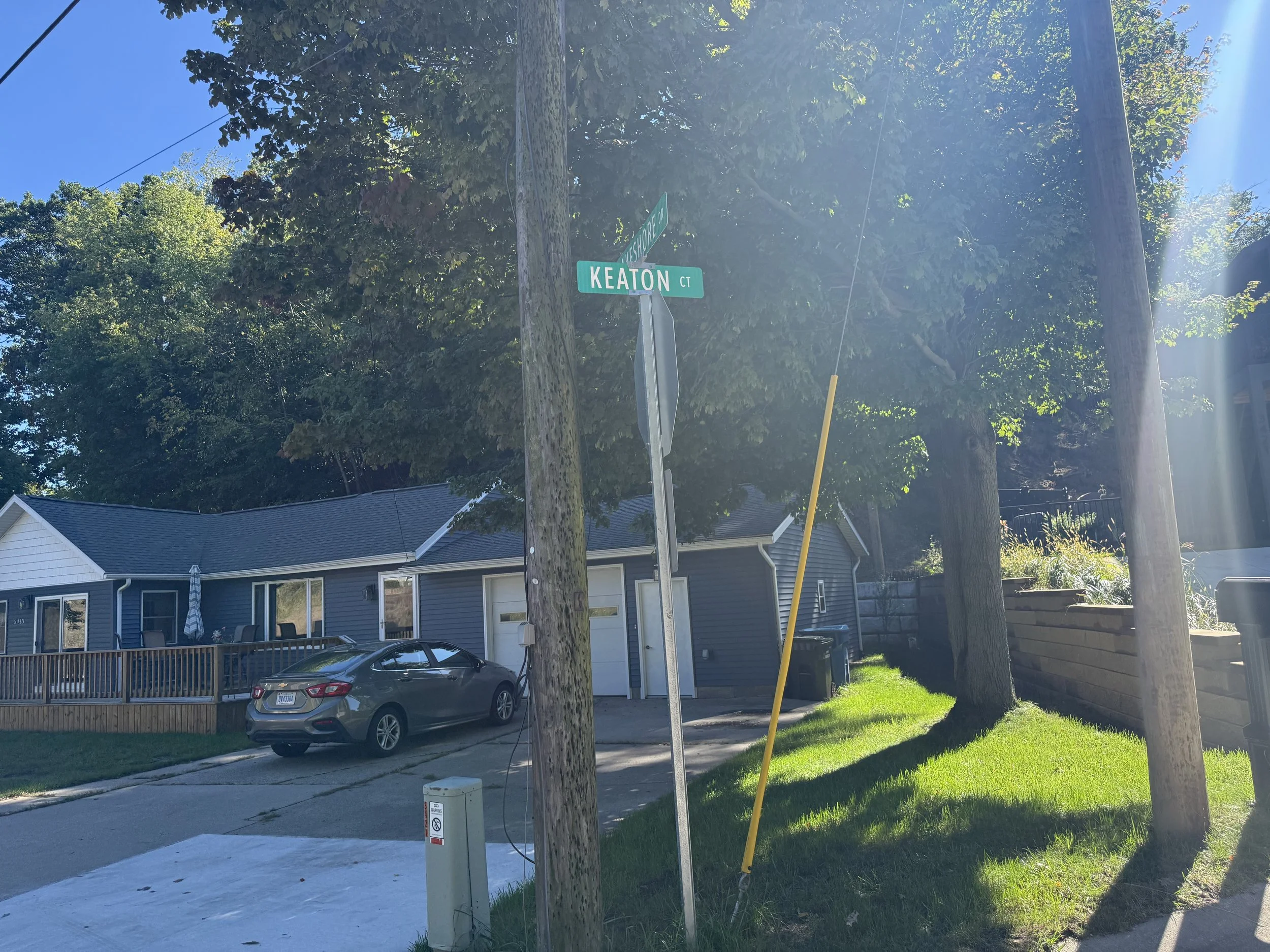 Street corner with street signs labeled "Keaton Ct" and "Lakeshore Dr." Residential houses and parked cars are visible in the background, with a large tree and a utility pole in the foreground. Clear blue sky overhead.